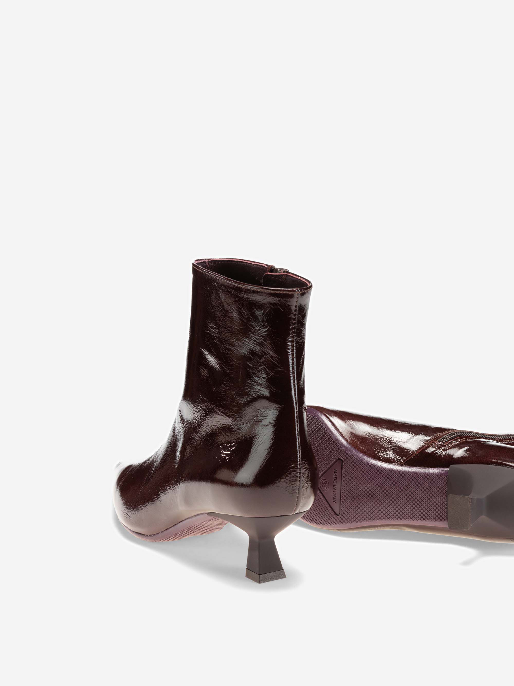 Zwei glänzende, weinrote Stiefeletten mit niedrigen, schrägen Absätzen. Ein Stiefel steht aufrecht, während der andere auf der Seite liegt und die Sohle und den Absatz vor einem schlichten weißen Hintergrund zeigt.