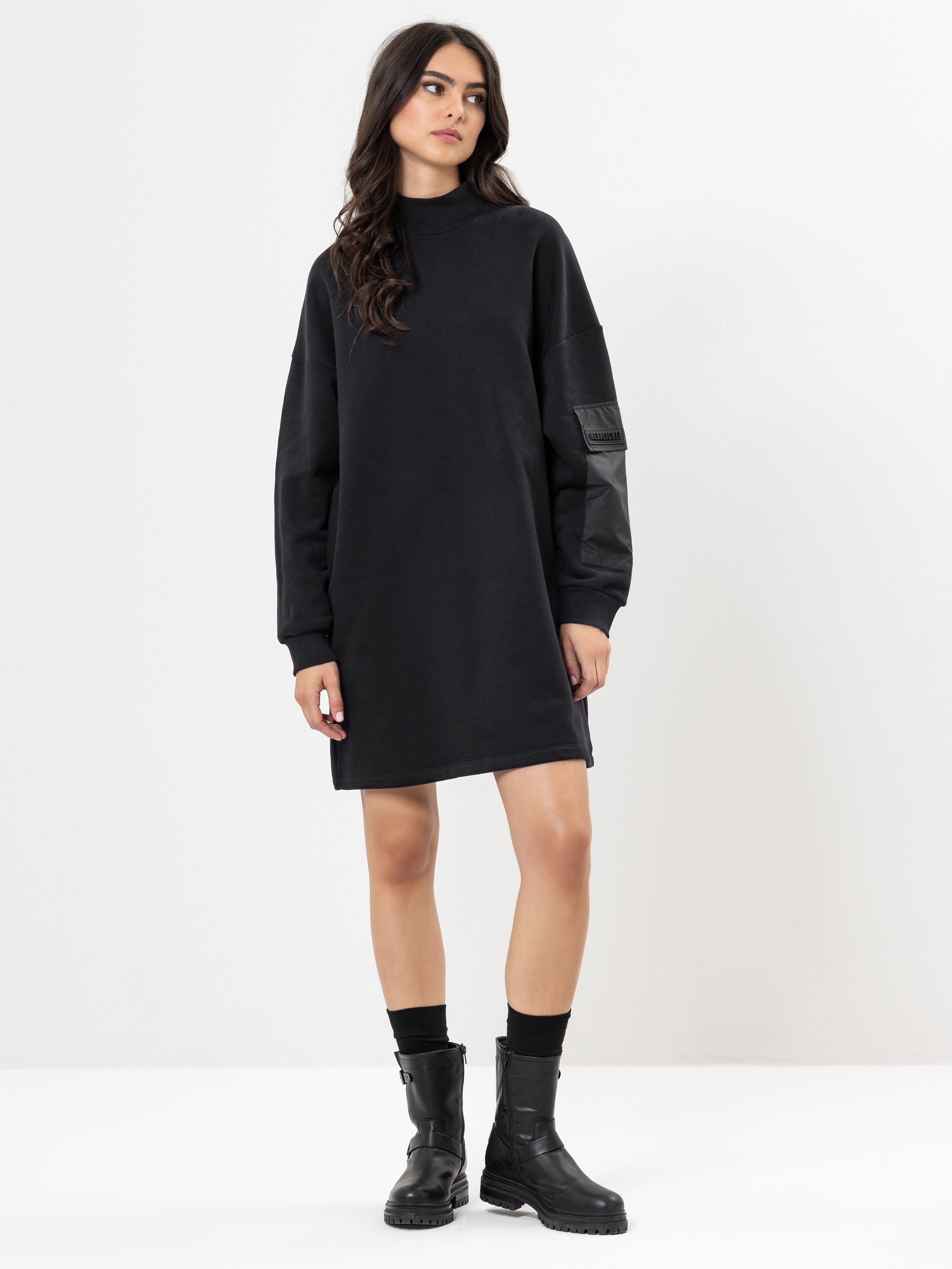 Eine Frau steht vor einem schlichten weißen Hintergrund und trägt ein locker sitzendes, schwarzes, langärmeliges Kleid mit hohem Ausschnitt und schwarze Ankle Boots. Ihr Haar ist gewellt und nach unten getragen, und sie hat einen neutralen Ausdruck.