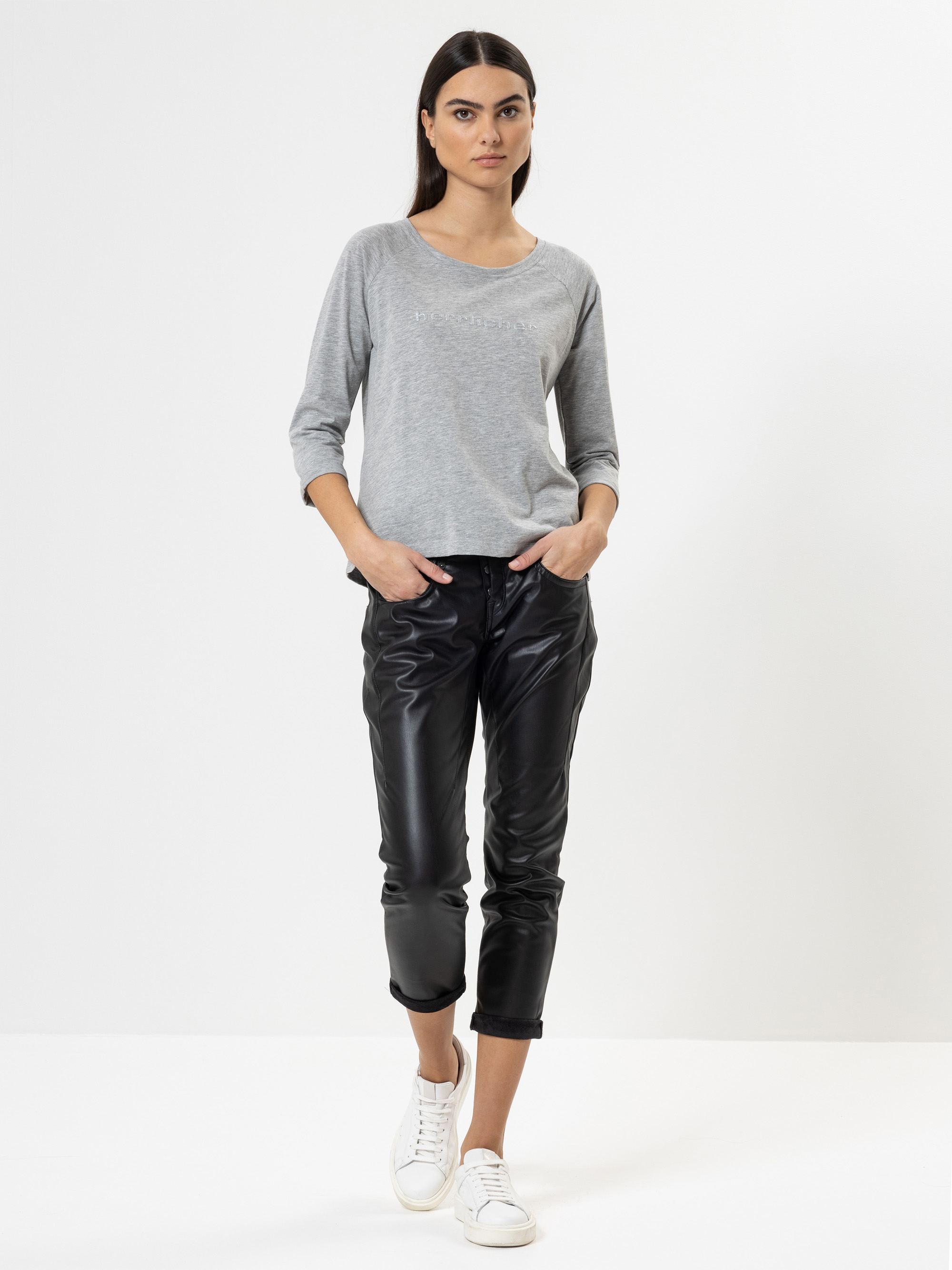 Eine Frau steht vor einem einfarbig weißen Hintergrund und trägt ein hellgraues, langärmeliges Oberteil, eine schwarze, glänzende, kurze Hose und weiße Turnschuhe. Sie hat langes dunkles Haar und einen neutralen Gesichtsausdruck, die Hände stecken i