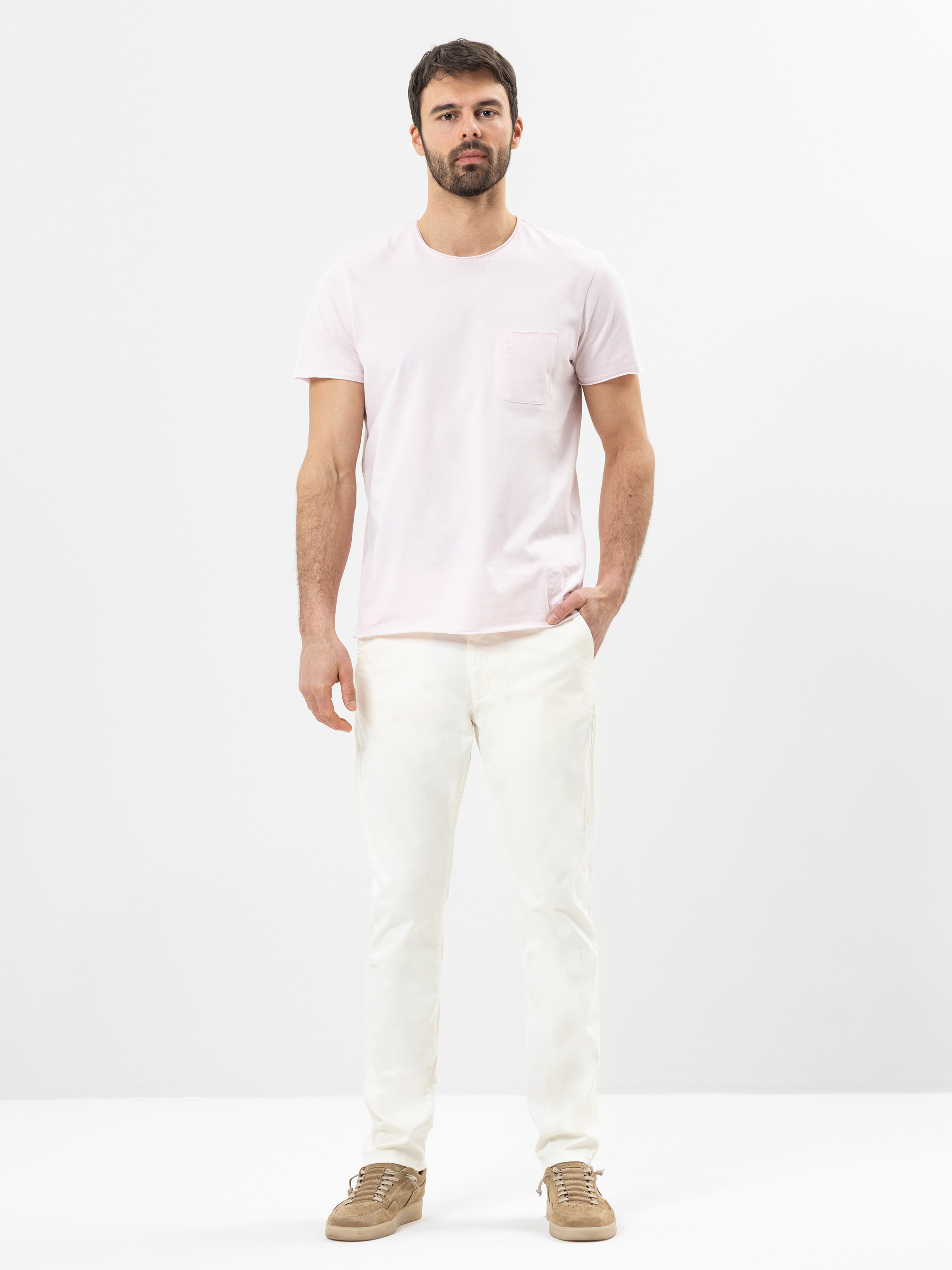 Ein Mann steht vor einem schlichten weißen Hintergrund und trägt ein hellrosa, kurzärmeliges T-Shirt mit einer Brusttasche, eine cremefarbene Hose und hellbraune Turnschuhe. Eine Hand steckt in seiner Hosentasche und er hat einen neutralen Gesichtsausd