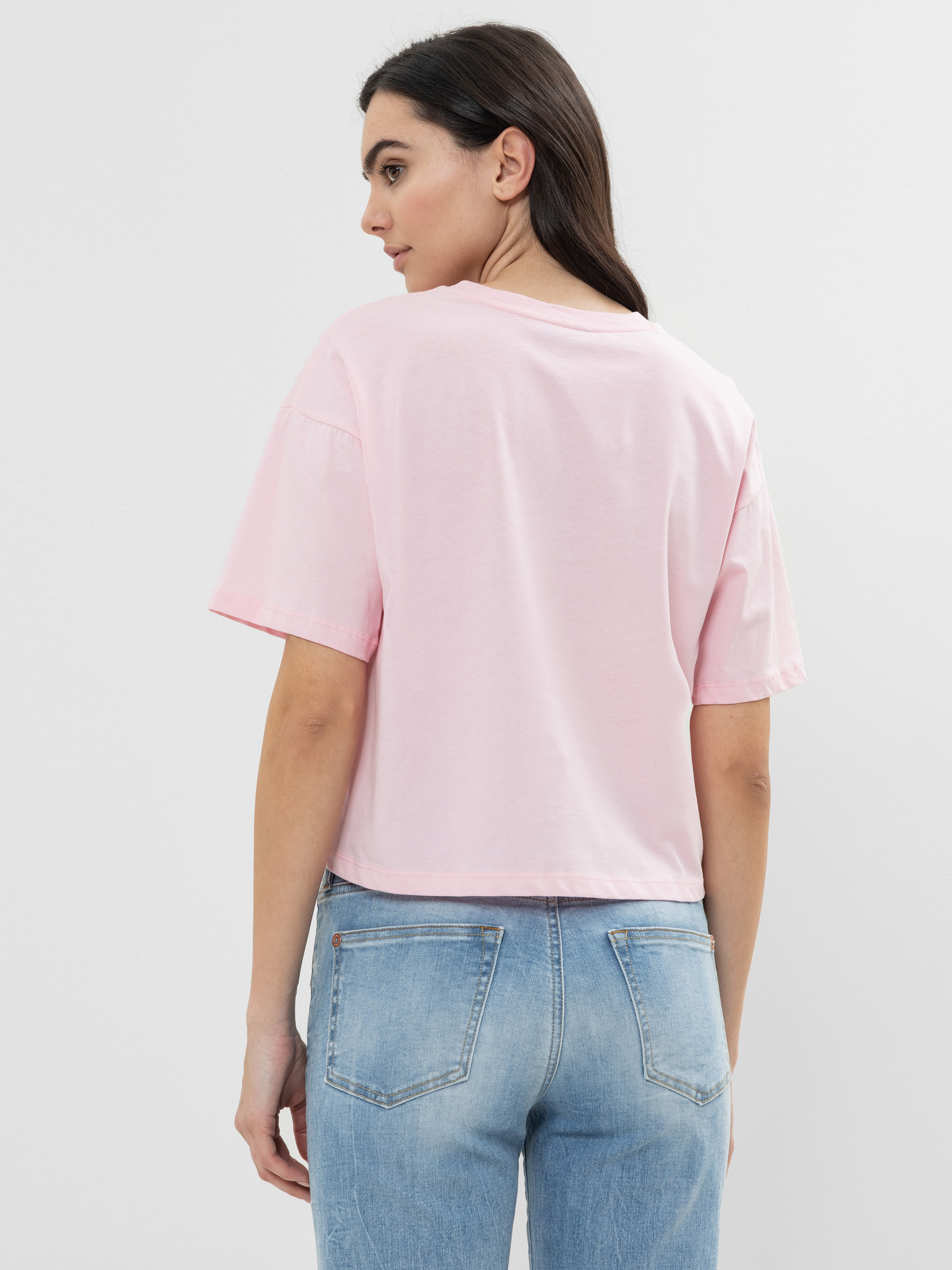 Eine Frau mit langen dunklen Haaren trägt ein hellrosa kurzärmeliges T-Shirt und hellblaue Jeans. Sie steht mit dem Gesicht zur Kamera und blickt über ihre linke Schulter vor einem einfarbigen Hintergrund.