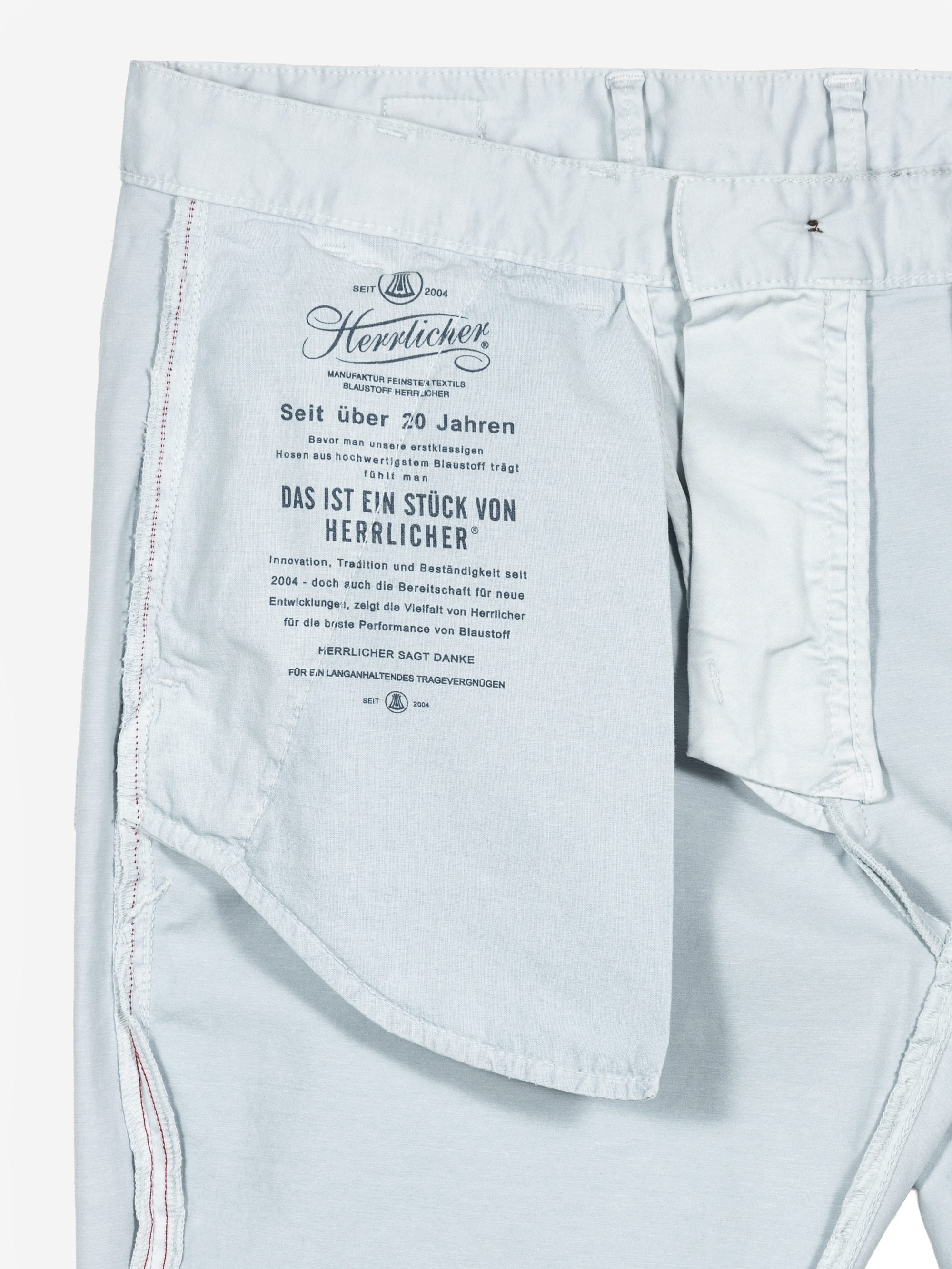 Die hellblaue Hose ist auf links gedreht und zeigt das Innenetikett mit schwarzer deutscher Schrift, dem Markennamen Herrlicher und roten Ziernähten entlang der Nähte. Das Etikett unterstreicht Qualität und Tradition.