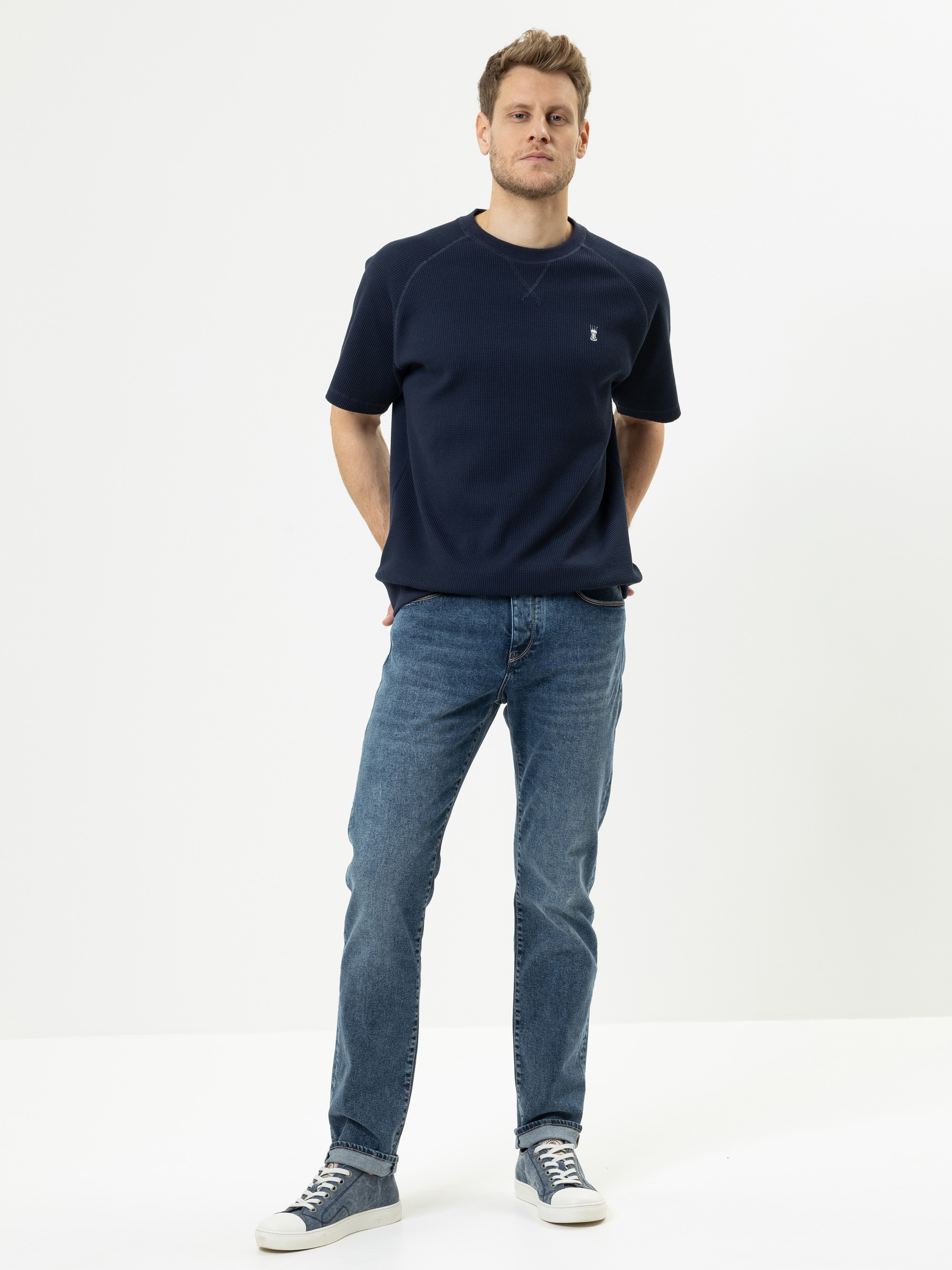 Eine Person steht vor einem schlichten weißen Hintergrund, trägt ein marineblaues, kurzärmeliges Hemd, blaue Jeans und graue Turnschuhe und hat die Hände auf dem Rücken.