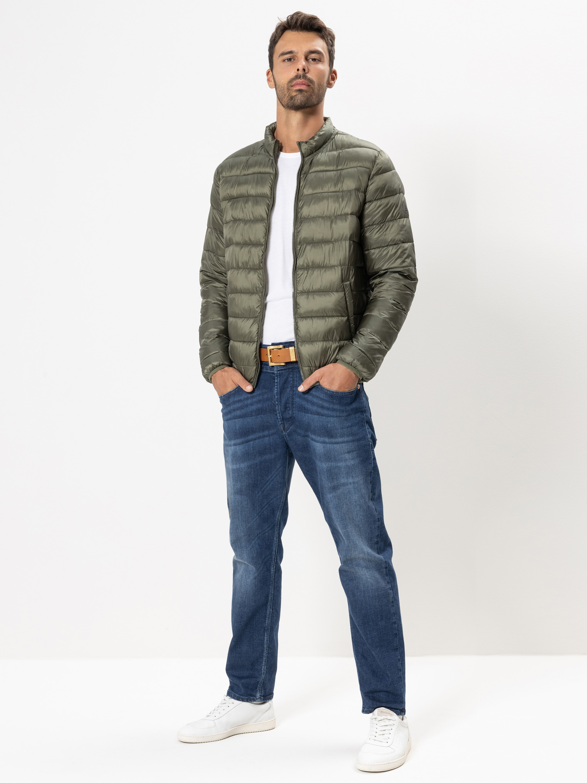 Ein Mann steht vor einem schlichten weißen Hintergrund, trägt eine grüne Pufferjacke, ein weißes T-Shirt, blaue Jeans, weiße Turnschuhe und einen hellbraunen Gürtel, die Hände in den Taschen und einen neutralen Blick.