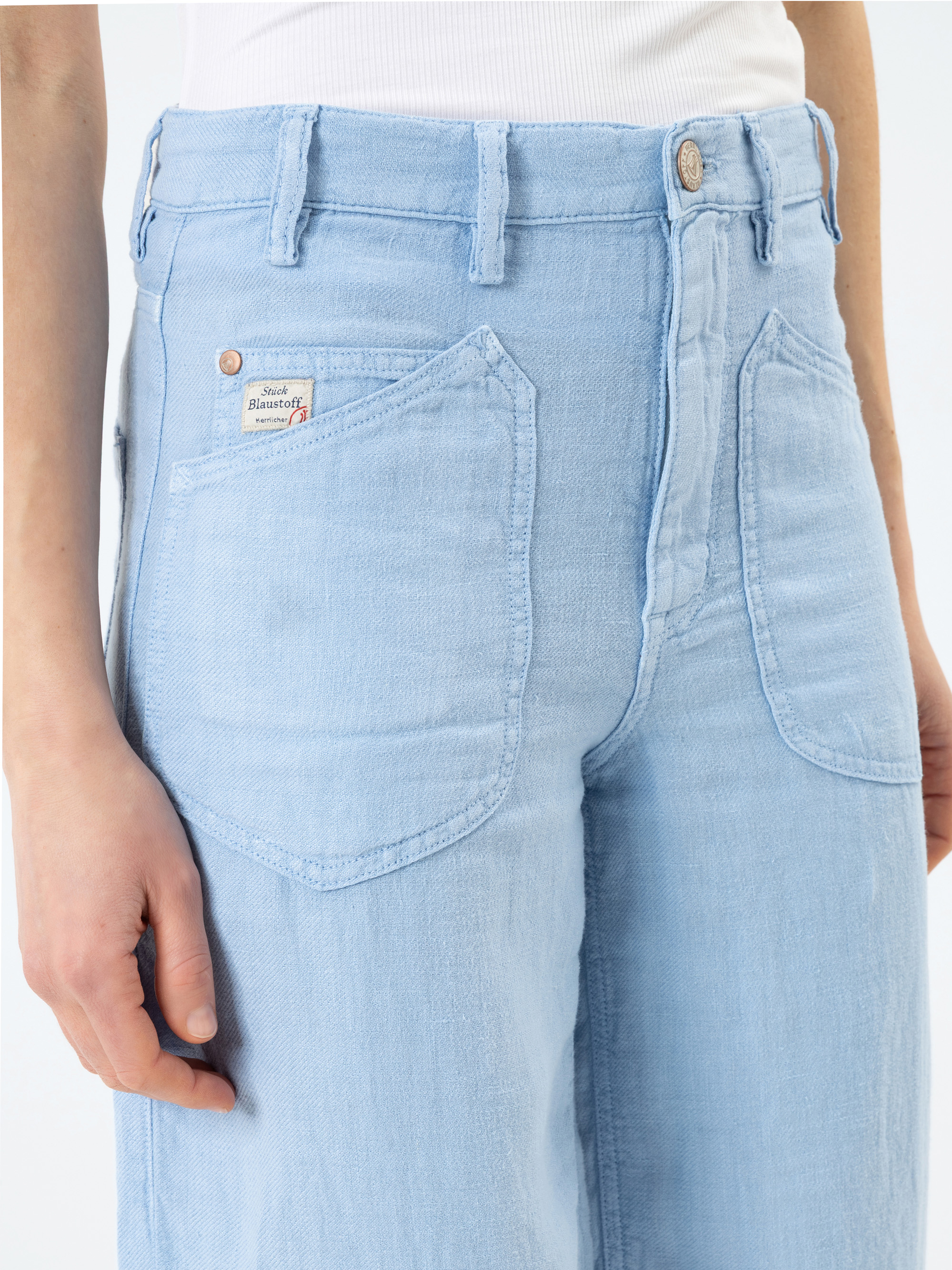 Eine Person trägt eine hellblaue Jeans mit hoher Taille und großen Vordertaschen und ein weißes Tanktop. Die Jeans hat ein kleines Etikett über der rechten Tasche. Nur der Torso und die Oberschenkel sind sichtbar.