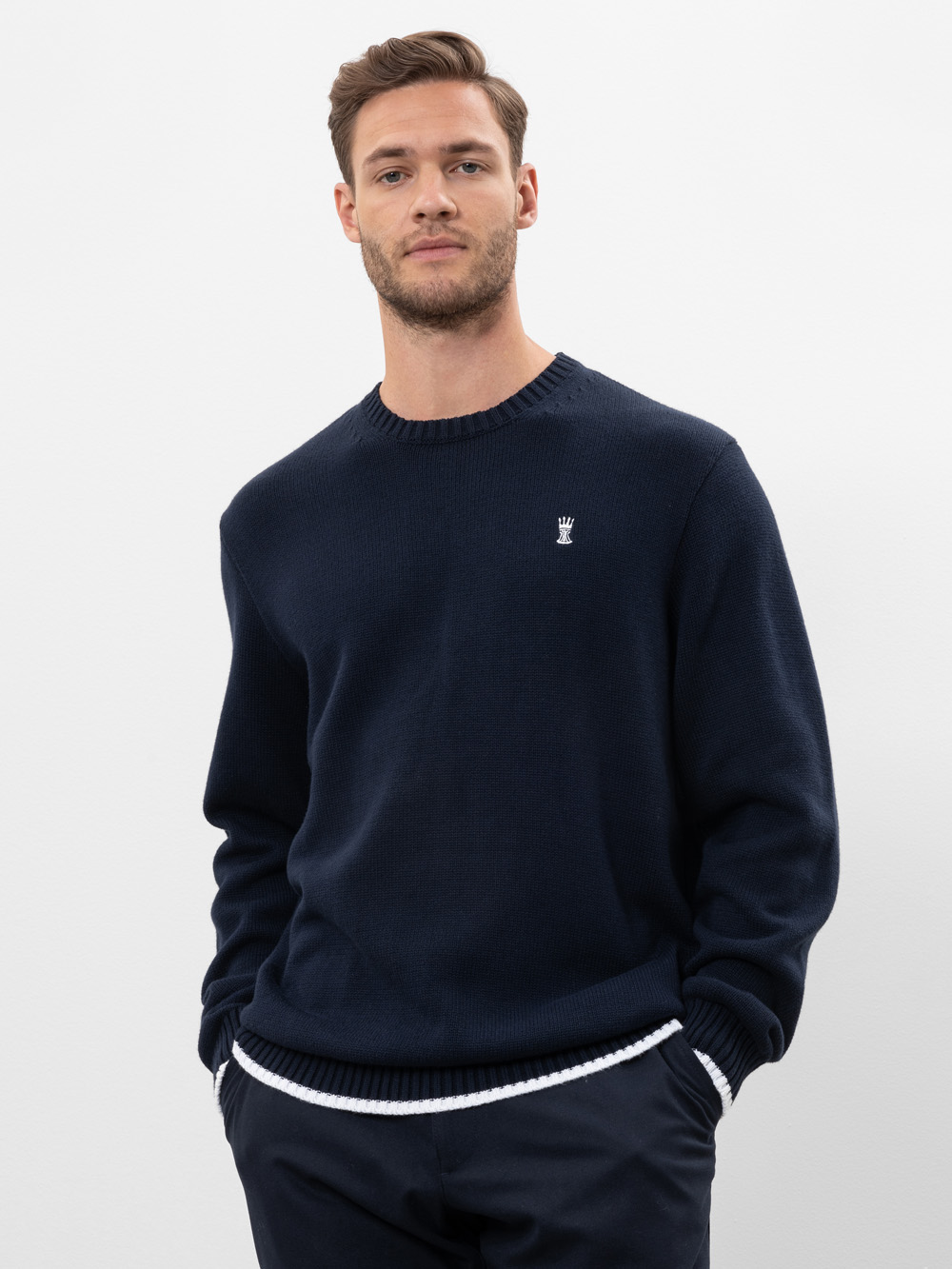 Ein Mann mit kurzen braunen Haaren und heller Gesichtsbehaarung trägt einen marineblauen Pullover mit einem kleinen weißen Kronenlogo auf der Brust, eine dunkle Hose und steht mit den Händen in den Taschen vor einem schlichten weißen Hintergrund.
