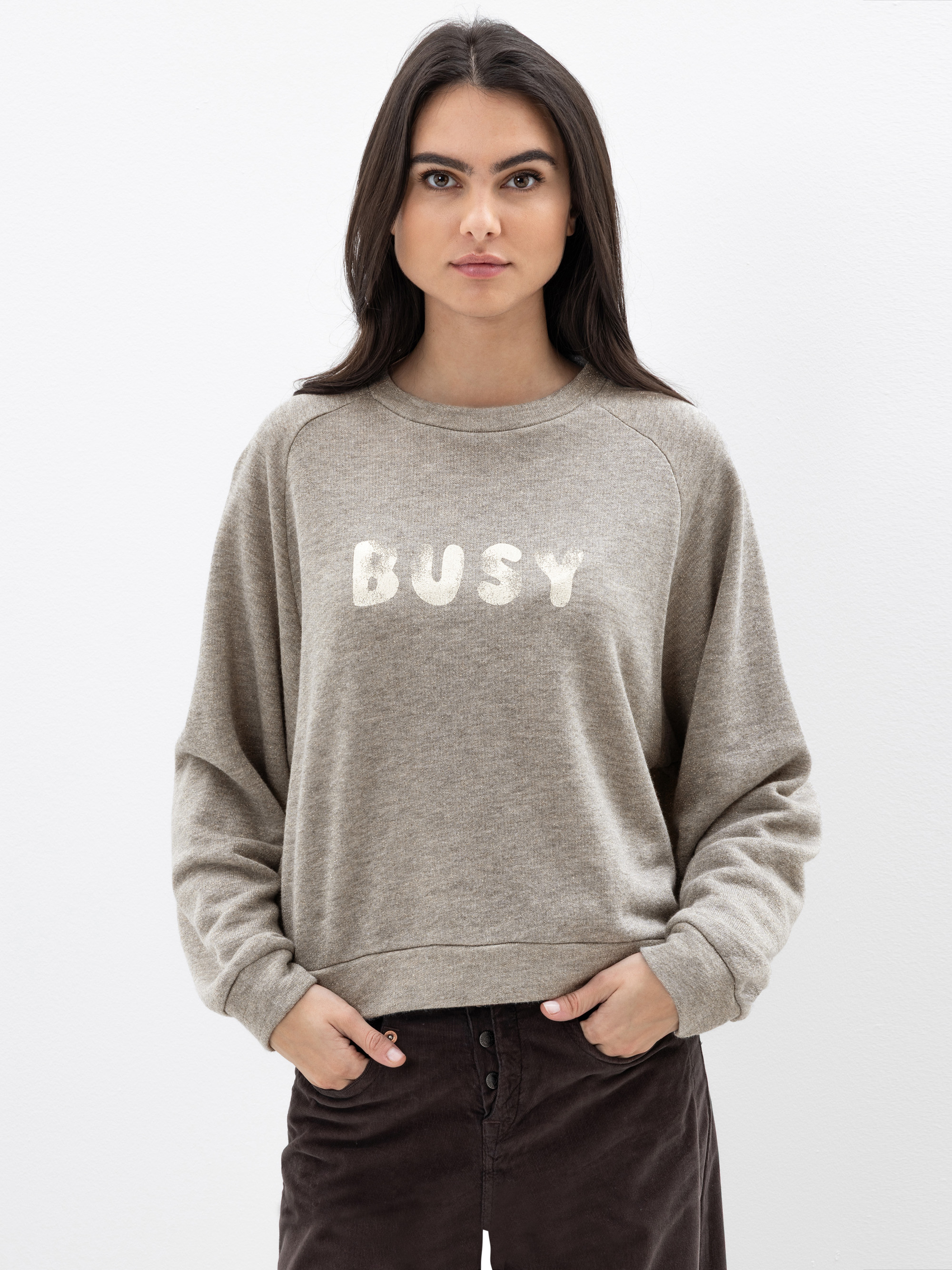 Eine Frau mit langen dunklen Haaren steht vor einem schlichten weißen Hintergrund. Sie trägt ein beiges Sweatshirt mit der Aufschrift BUSY auf der Vorderseite und eine dunkelbraune Hose. Sie hat einen neutralen Gesichtsausdruck und ihre Hände stecken i