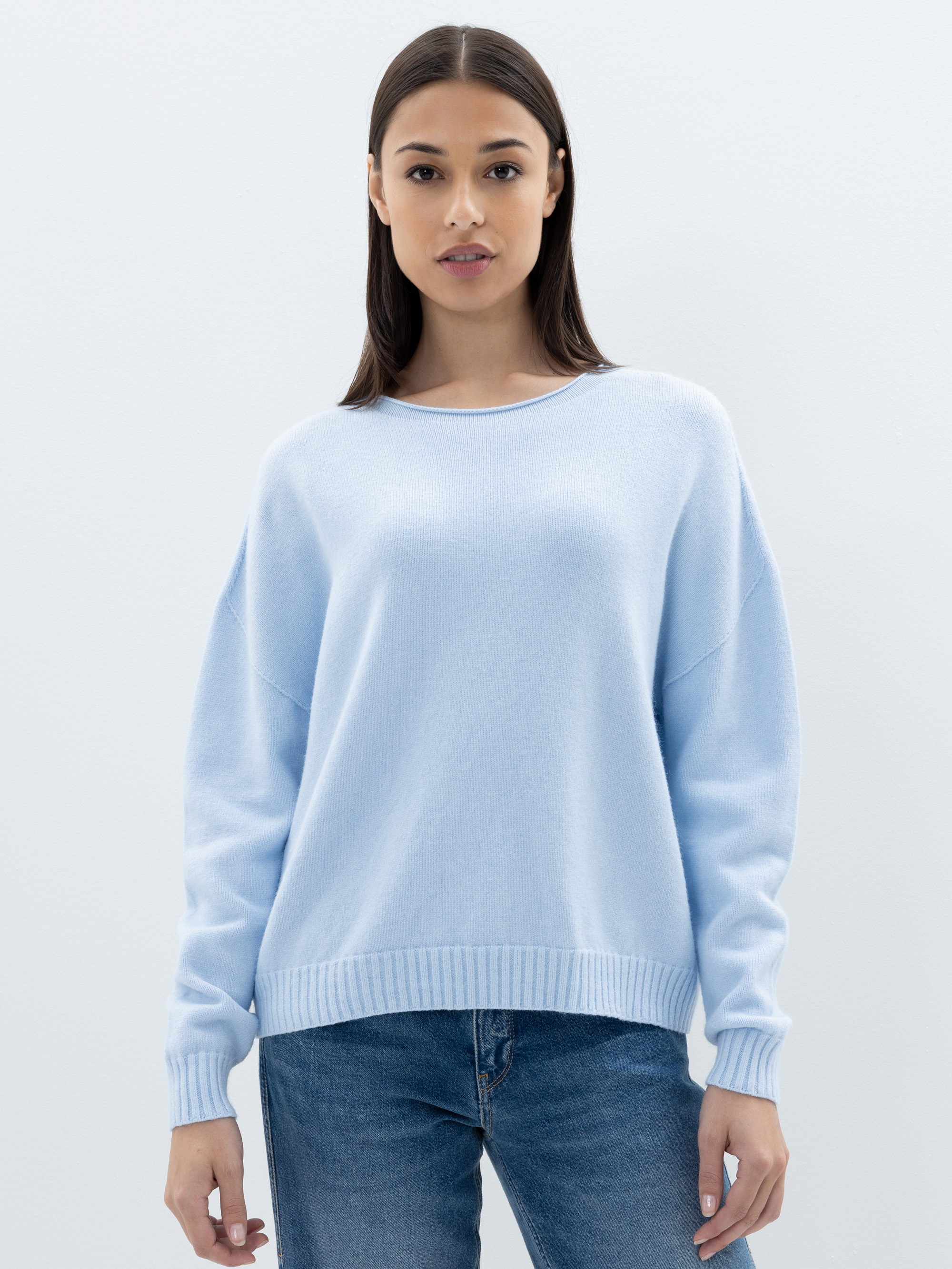 Eine Frau mit glattem braunem Haar, die einen hellblauen Pullover und blaue Jeans trägt, steht mit dem Gesicht zur Kamera vor einem schlichten weißen Hintergrund.