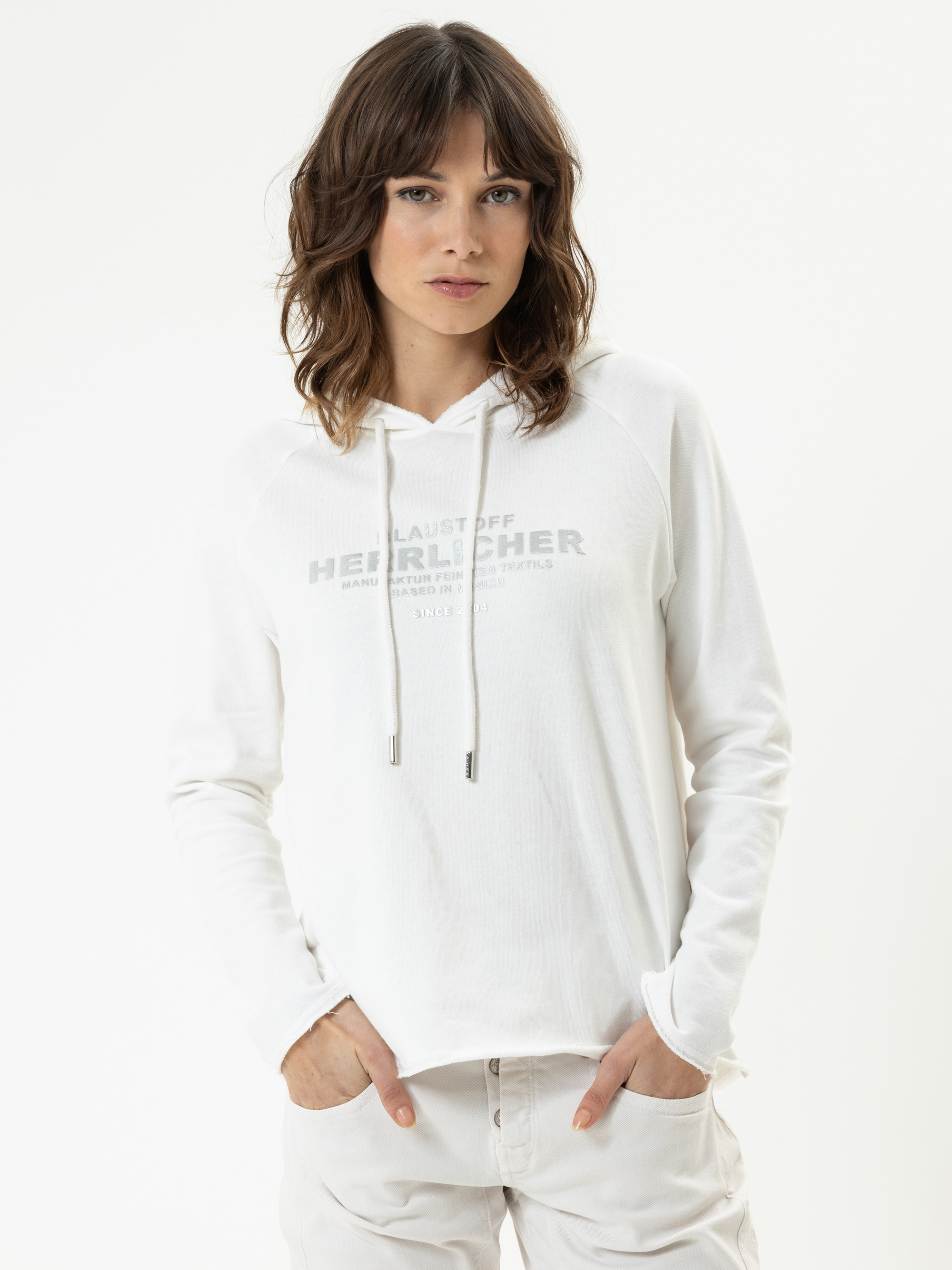 Eine Frau mit gewelltem braunem Haar trägt ein weißes Kapuzensweatshirt mit schwacher Aufschrift auf der Vorderseite und eine helle Hose. Sie steht mit den Händen in den Taschen vor einem schlichten weißen Hintergrund.
