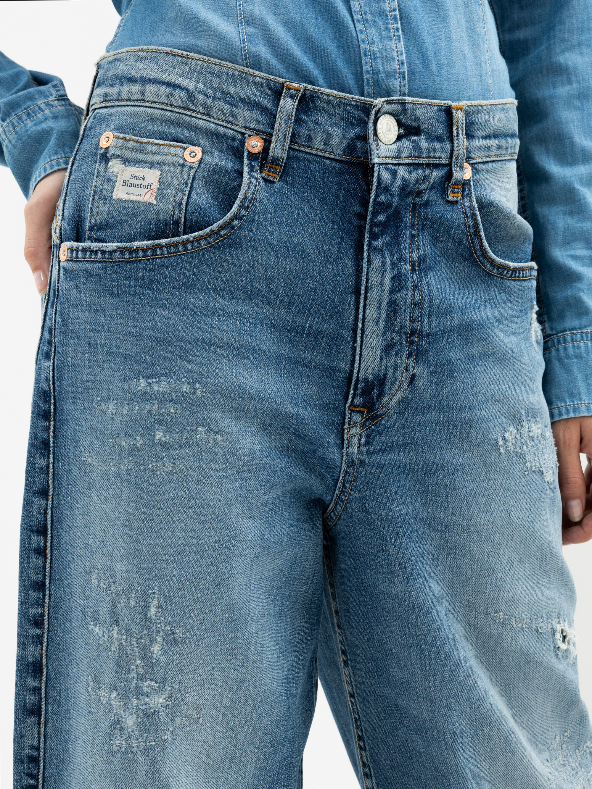 Nahaufnahme einer Person, die eine verblichene blaue Jeans und ein dazu passendes Jeanshemd trägt und eine Hand in die Jeanstasche gesteckt hat. Die Jeans trägt ein kleines Etikett über der Tasche und hat eine leicht abgenutzte Struktur.