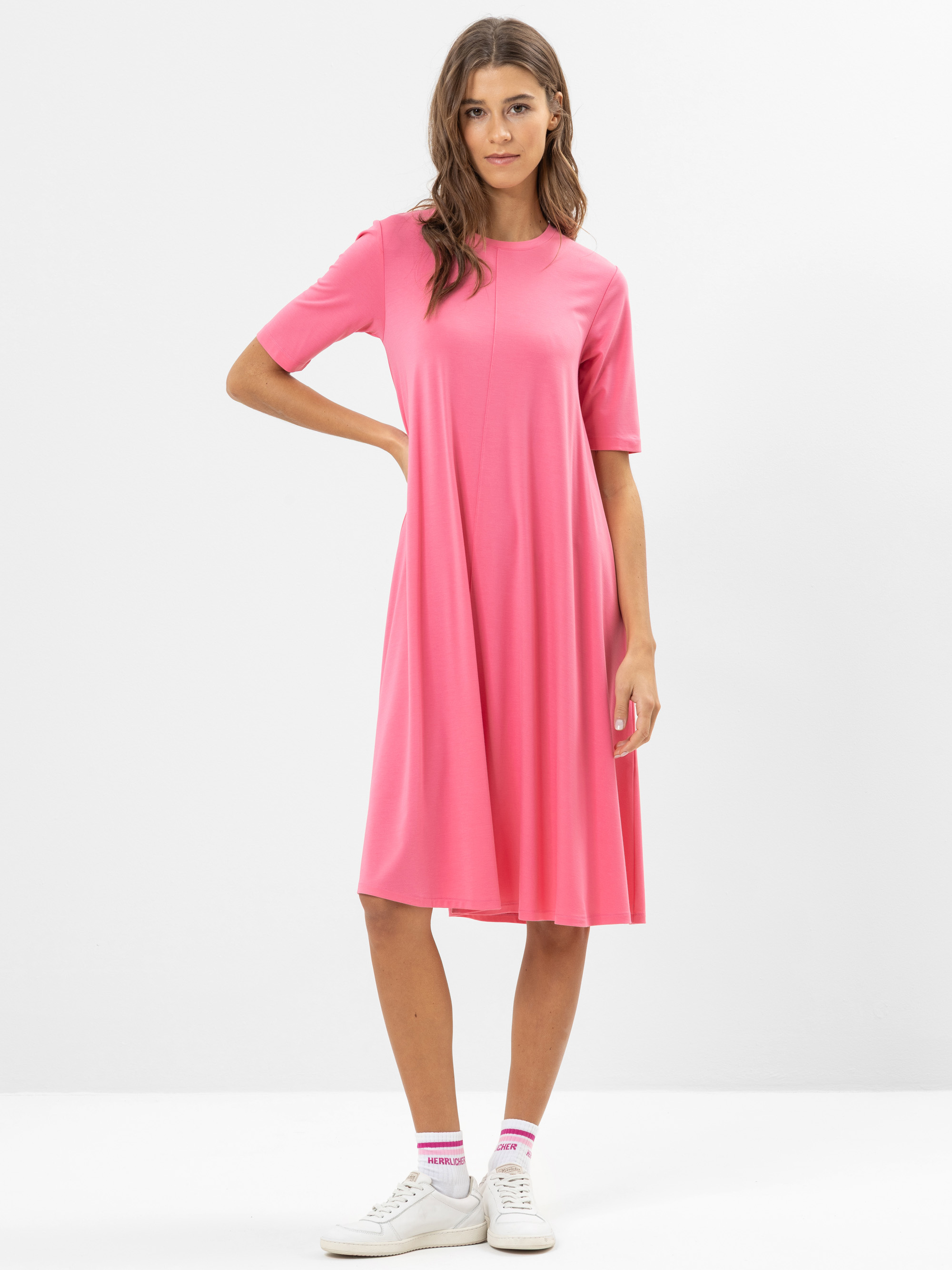 Eine Frau steht vor einem schlichten weißen Hintergrund und trägt ein knielanges, kurzärmeliges, rosafarbenes Kleid, gepaart mit weißen Turnschuhen und weißen Socken mit rosa Akzenten. Sie hat langes braunes Haar und eine entspannte, selbstbewusste P