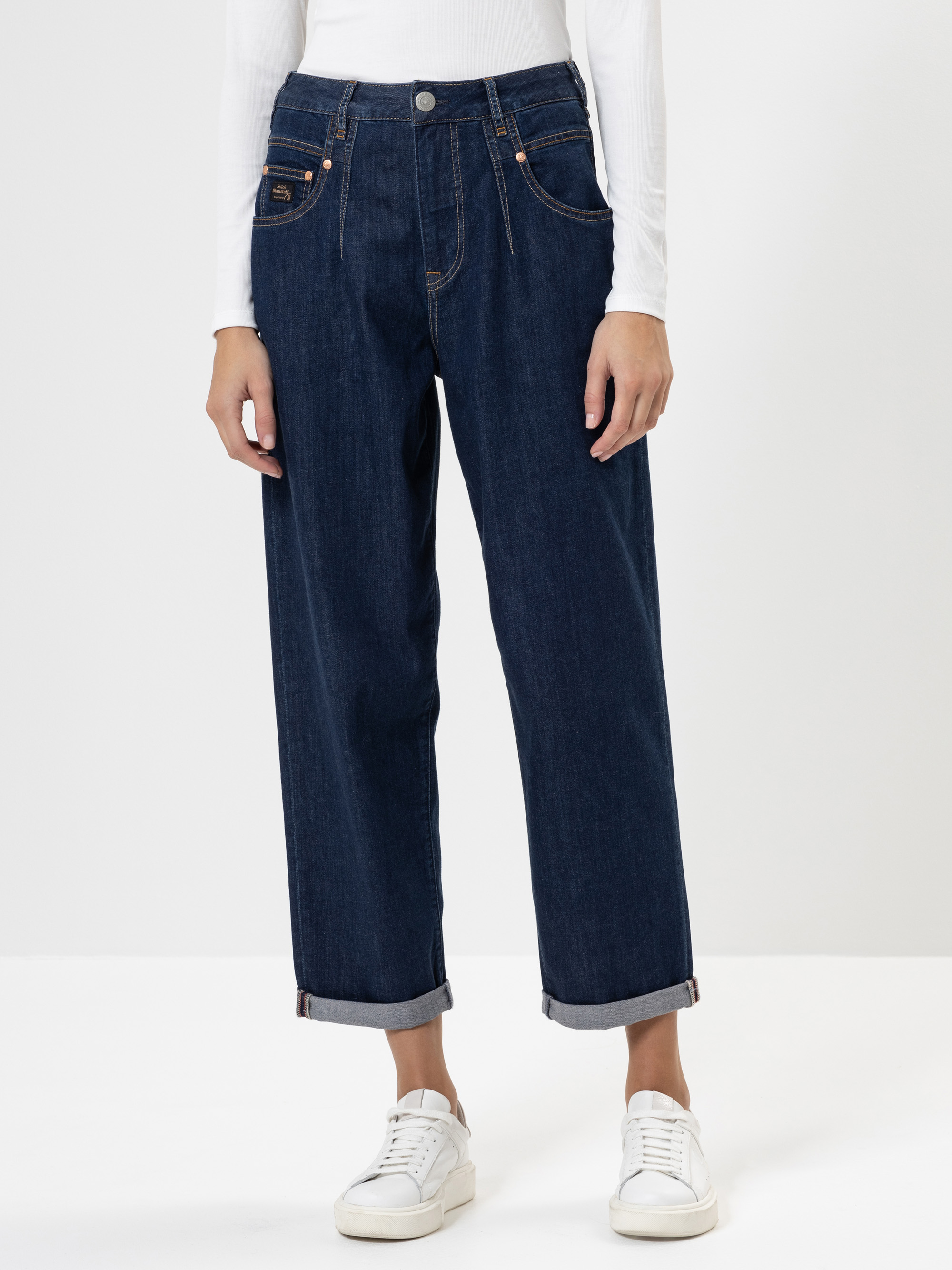 Eine Person, die dunkelblaue, weit geschnittene Jeans, ein weißes, langärmeliges Oberteil und weiße Turnschuhe trägt, steht vor einem schlichten weißen Hintergrund. Die Jeans sind an den Knöcheln mit Bündchen versehen.