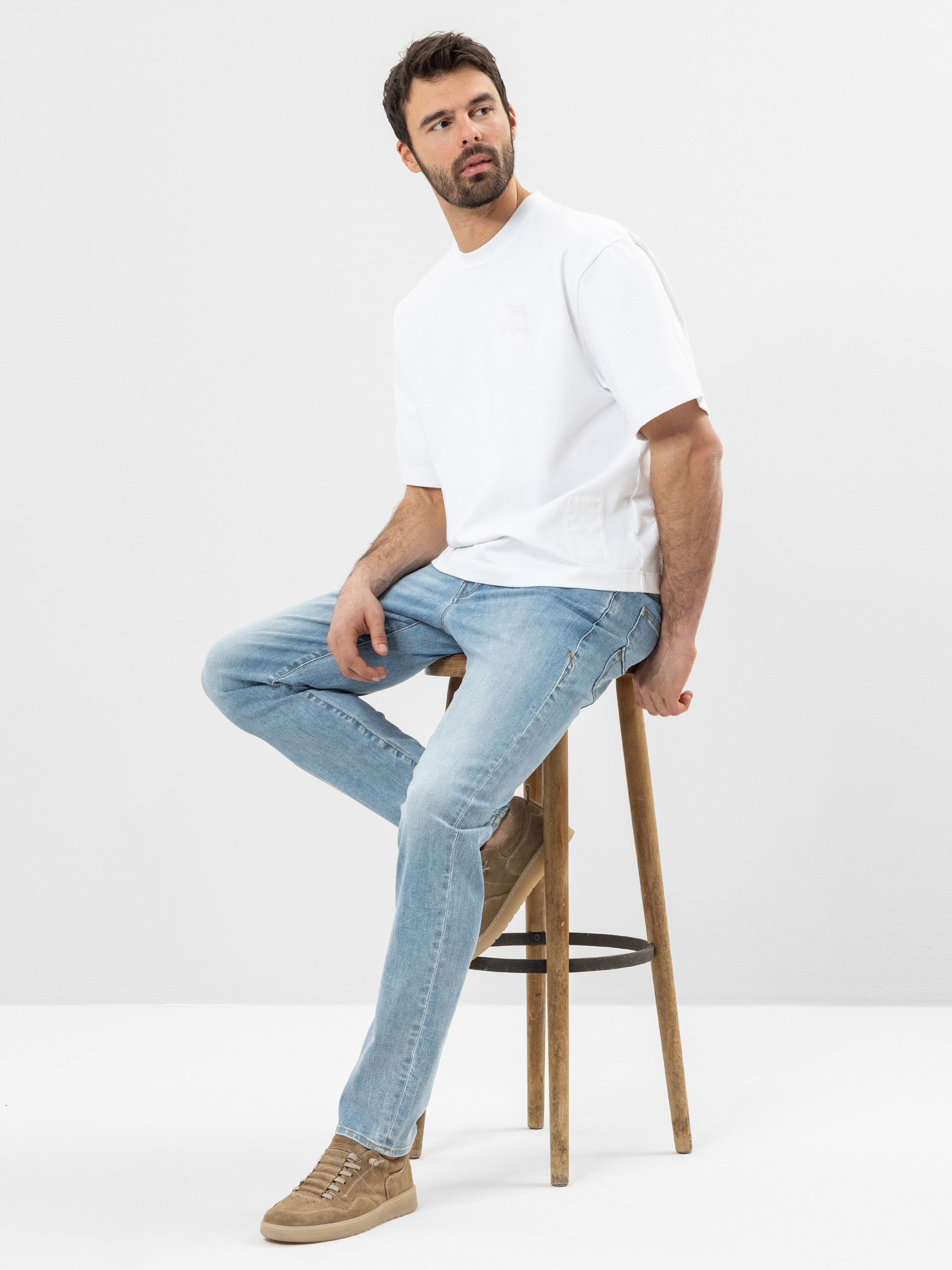 Ein Mann mit dunklem Haar und Bart sitzt auf einem Holzhocker vor einem schlichten weißen Hintergrund und trägt ein weißes T-Shirt, hellblaue Jeans und hellbraune Turnschuhe. Er blickt mit neutraler Miene nach links.