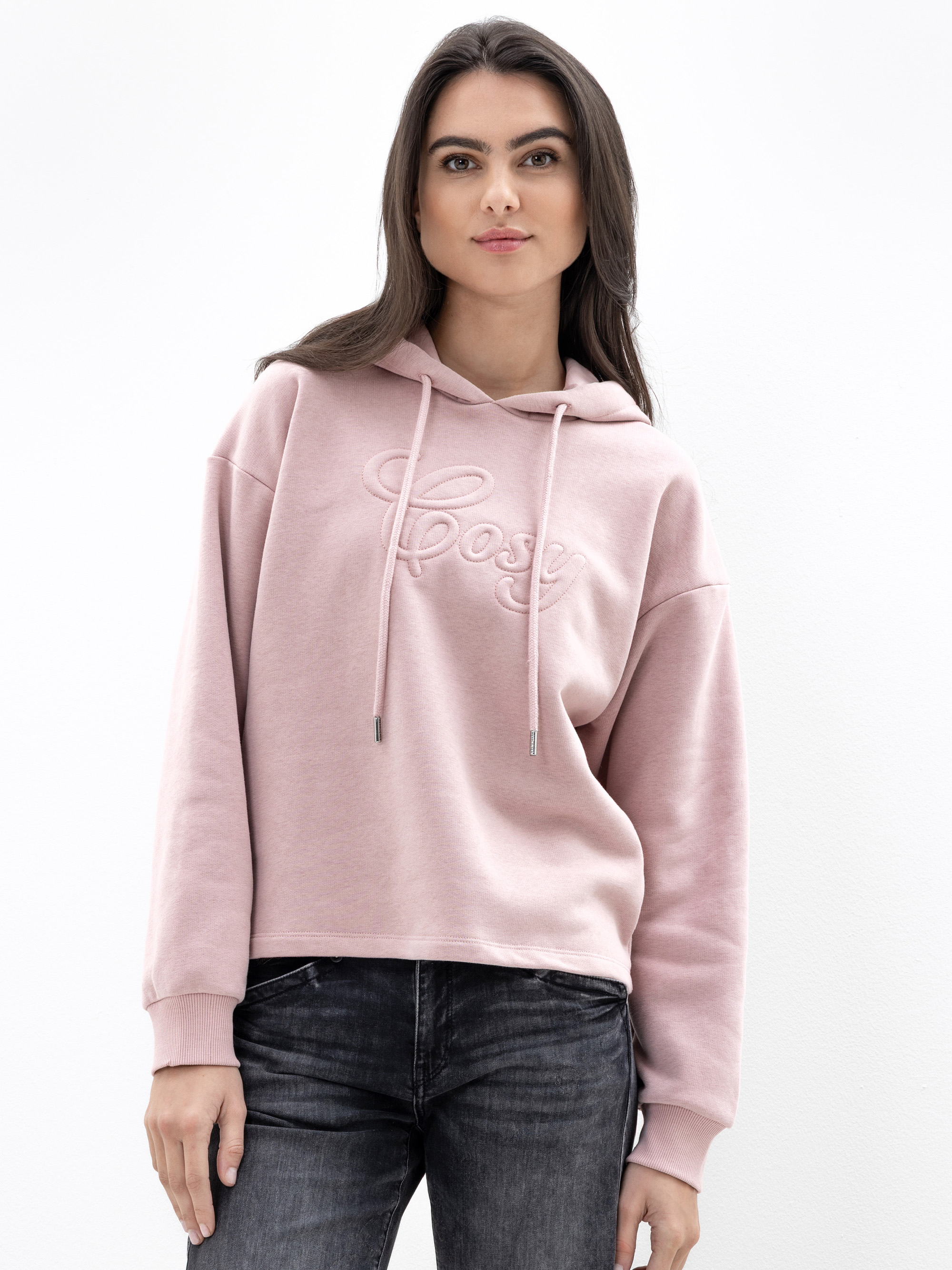 Eine Frau mit langen dunklen Haaren trägt einen hellrosa Kapuzenpulli mit dem aufgestickten Wort Cosy auf der Vorderseite und eine dunkle Jeans, die vor einem schlichten weißen Hintergrund steht.