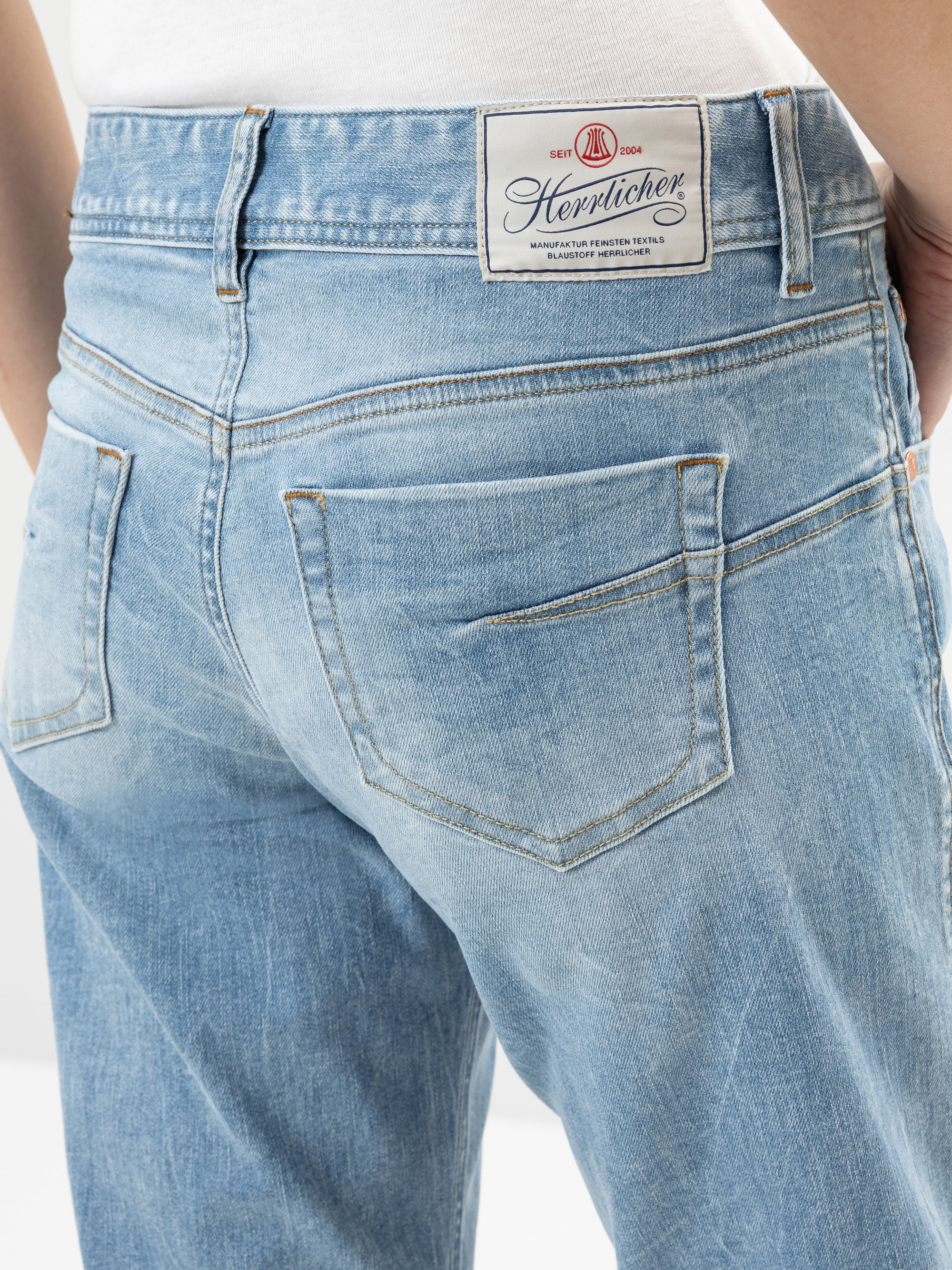 Nahaufnahme einer Person, die eine hellblaue Denim-Jeans trägt, mit Gesäßtaschen und einem weißen Aufnäher mit der Marke Herrlicher am Bund.