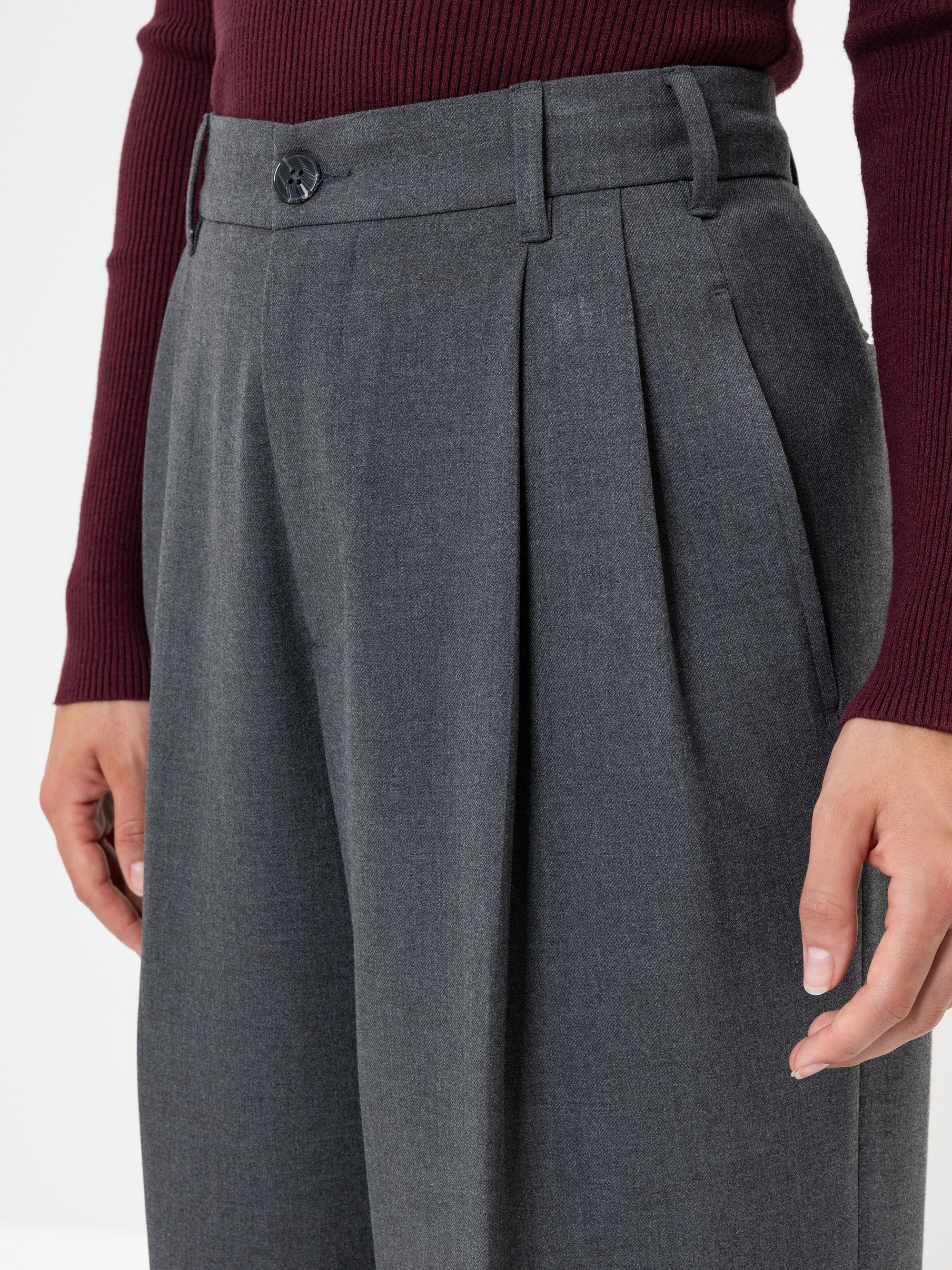 Eine Person, die eine graue Hose mit hoher Taille und Plisseefalten und ein langärmeliges weinrotes Oberteil trägt, wird von der Taille abwärts mit entspannten Händen an der Seite gezeigt.