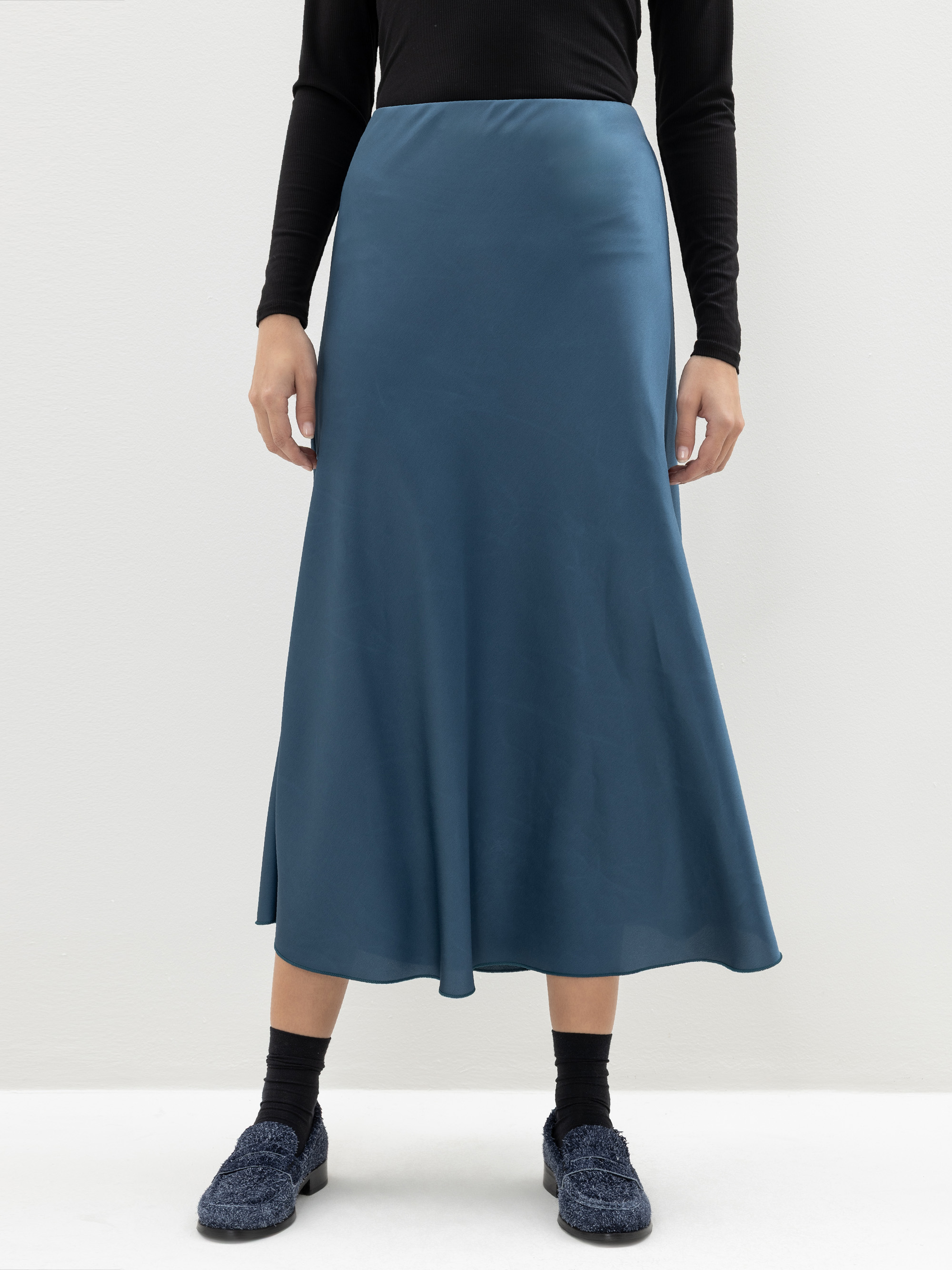 Eine Person, die ein schwarzes langärmeliges Oberteil, einen weinroten Midi-Satinrock, schwarze Socken und blaue strukturierte Schuhe trägt, steht vor einem schlichten hellen Hintergrund. Ihr Kopf ist auf dem Bild nicht zu sehen.