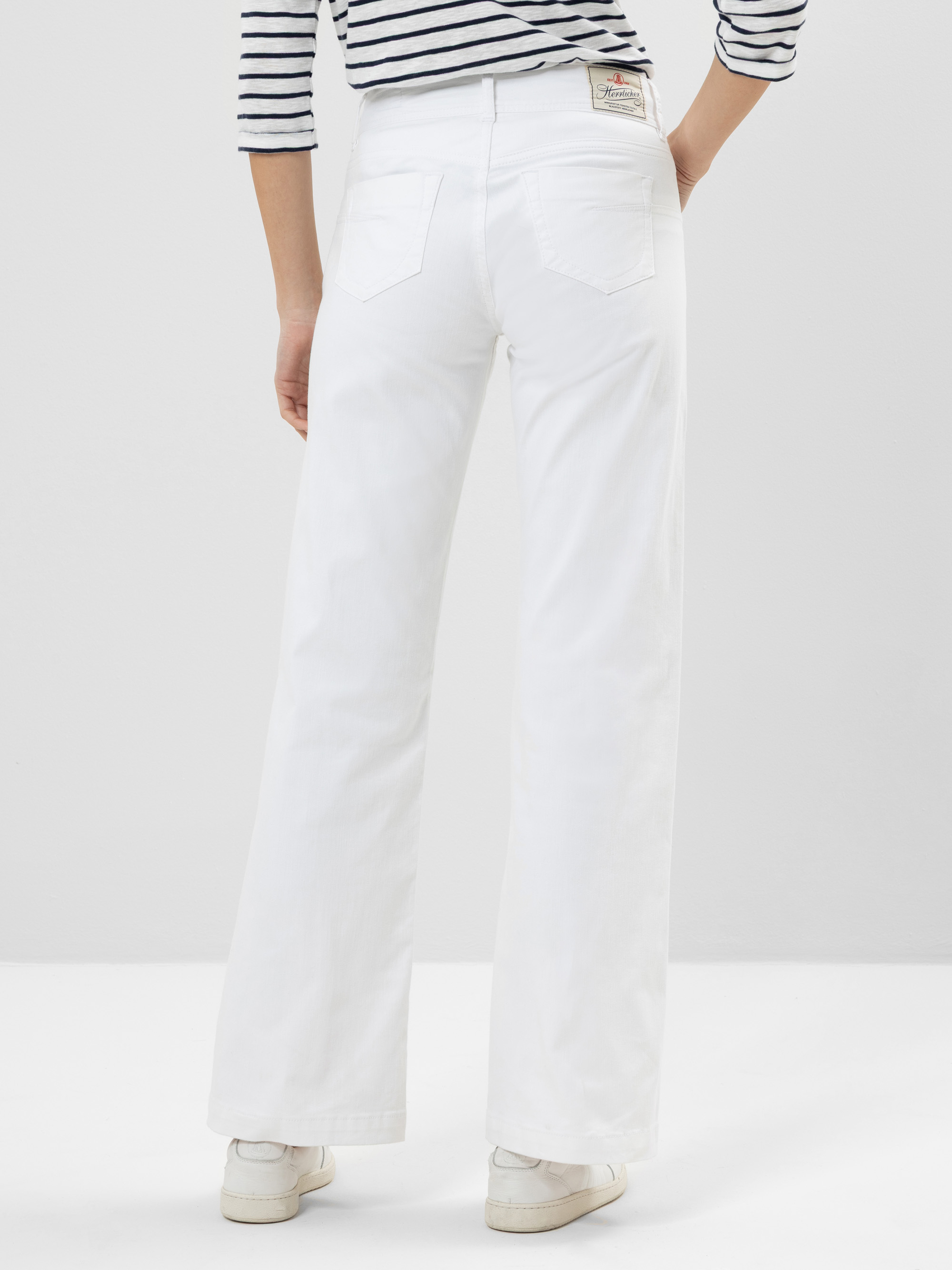 Eine Person steht mit dem Gesicht zur Wand und trägt ein schwarz-weiß gestreiftes langärmeliges Hemd, weiße Jeans mit hoher Taille und weiten Beinen sowie weiße Turnschuhe vor einem schlichten hellen Hintergrund.
