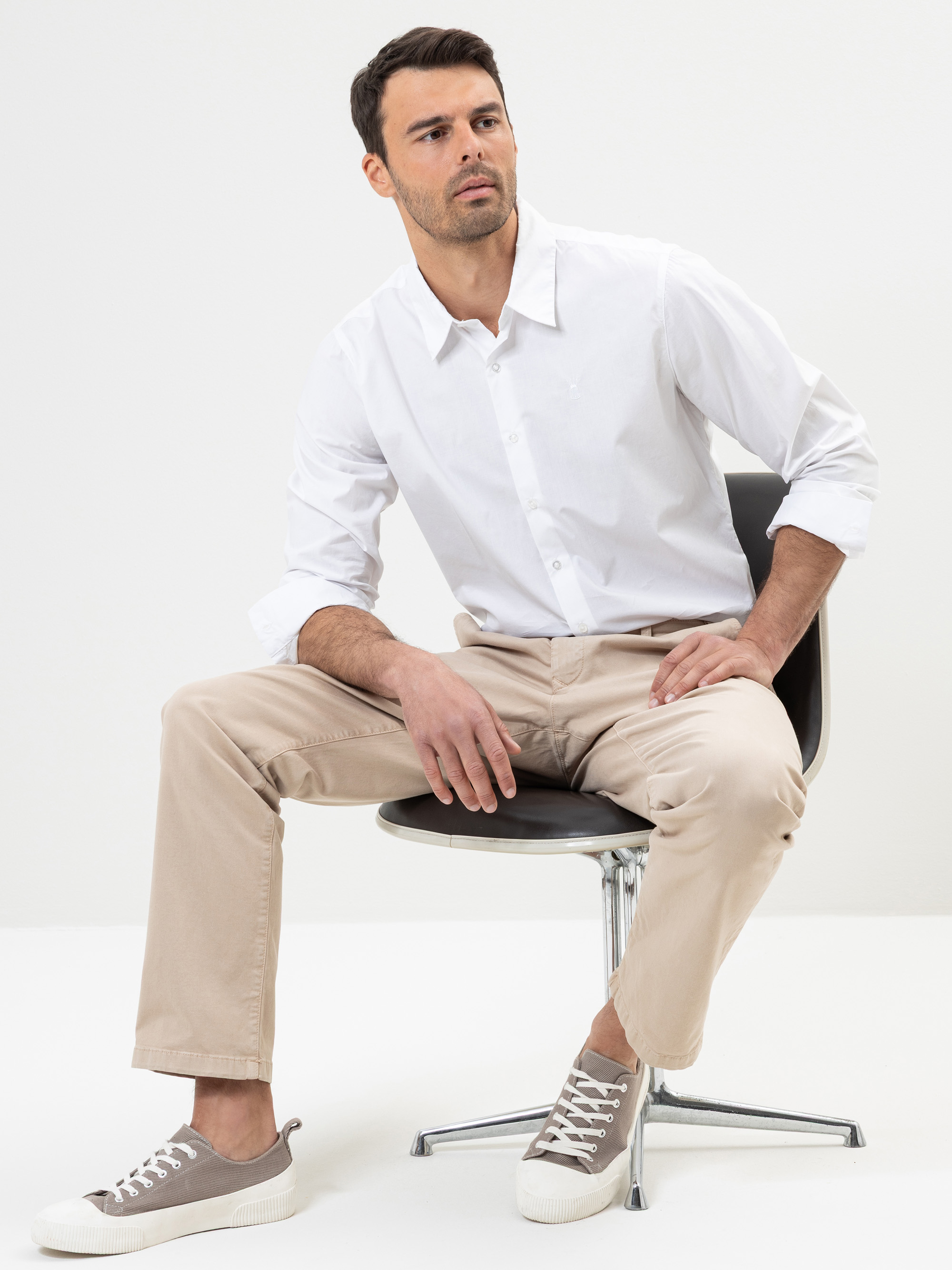 Ein Mann mit kurzen dunklen Haaren, der ein weißes Hemd, eine beigefarbene Hose und graue Turnschuhe trägt, sitzt auf einem schwarzen Drehstuhl vor einem schlichten weißen Hintergrund und schaut leicht nach links.