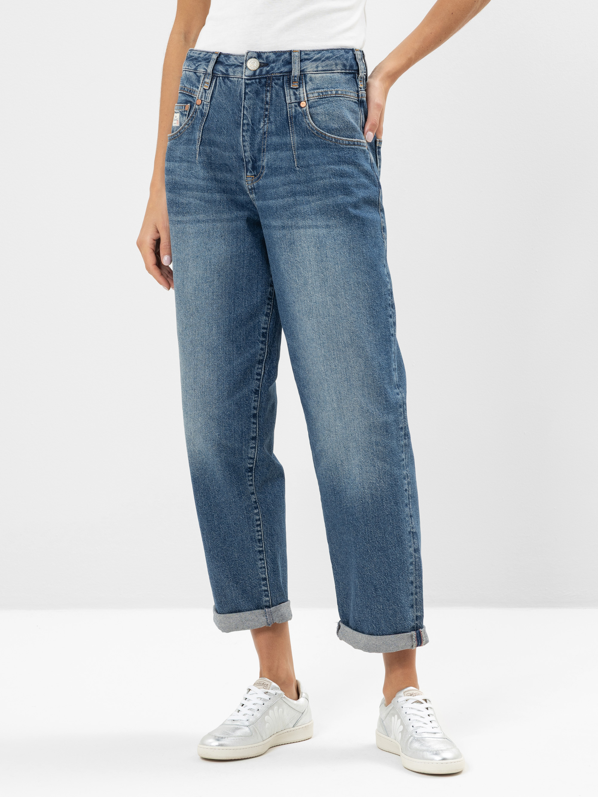 Eine Person, die ein weißes Hemd, blaue Jeans mit geradem Bein und silberne Turnschuhe trägt, steht vor einem schlichten weißen Hintergrund und hat eine Hand in der Tasche.