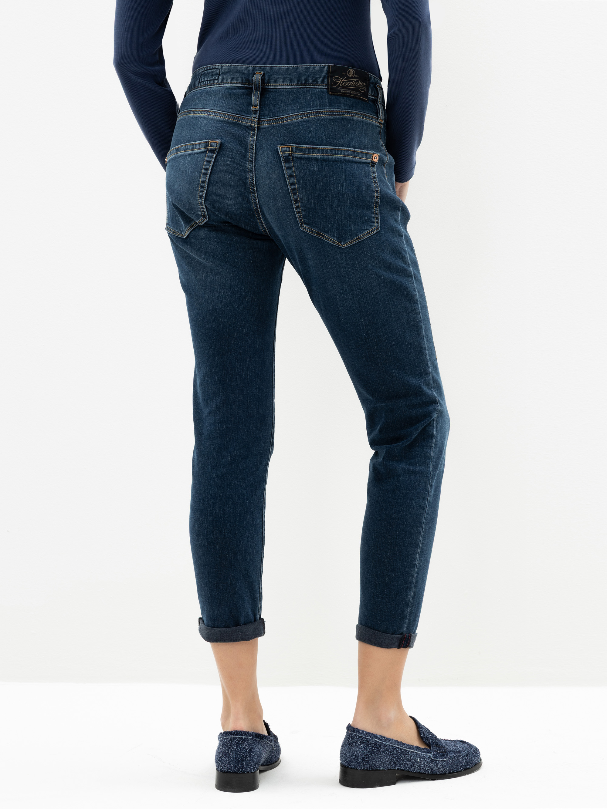 Eine Person, die mit dem Rücken zur Kamera steht, trägt dunkelblaue Jeans mit Bündchen, ein marineblaues langärmeliges Oberteil und blaue Slipper mit Struktur vor einem schlichten weißen Hintergrund.