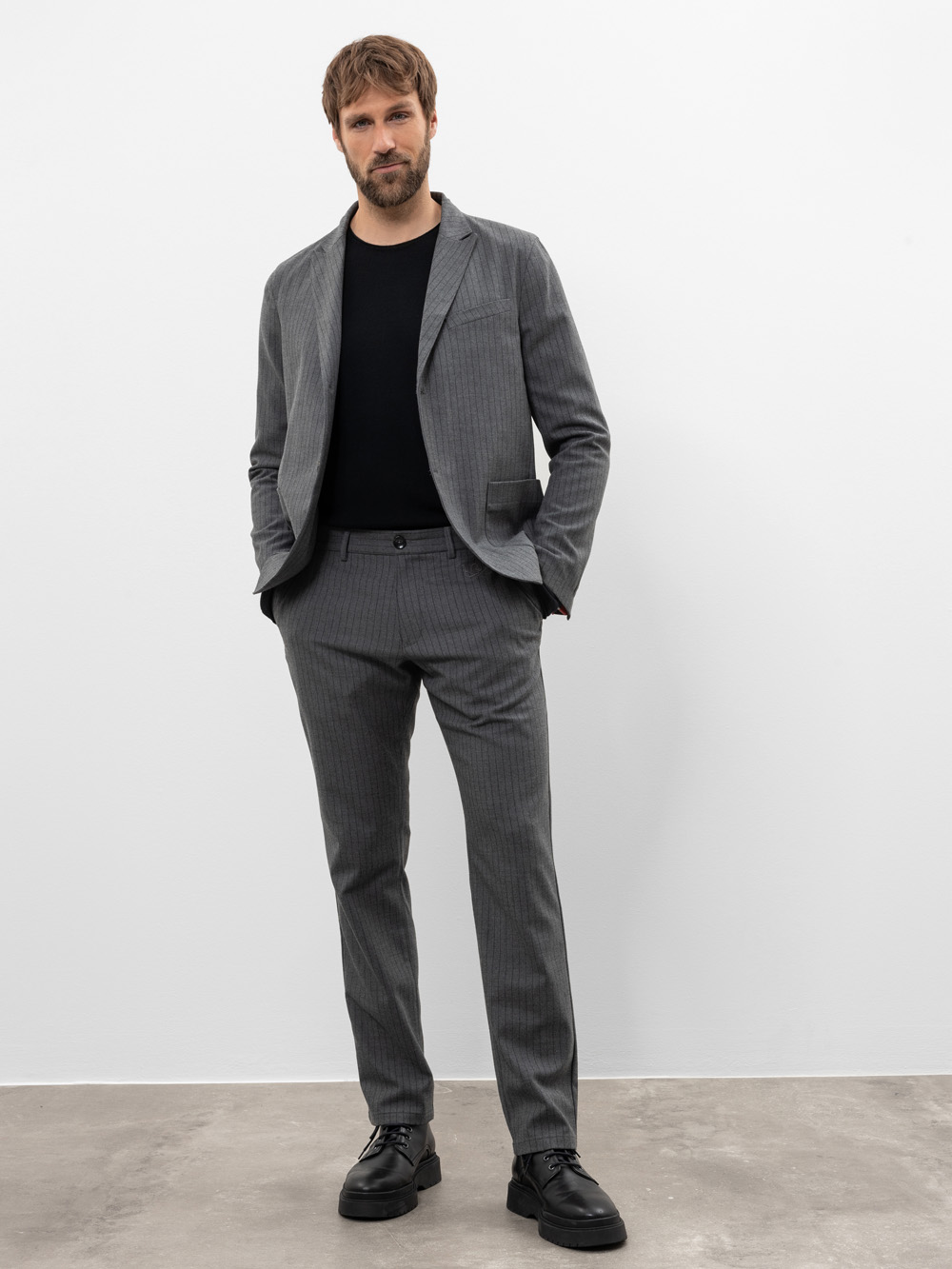 Ein Mann mit kurzen Haaren und Bart steht vor einer schlichten weißen Wand. Er trägt einen grauen Nadelstreifenanzug mit einem schwarzen Hemd und schwarzen Schuhen. Er hat eine Hand in der Tasche und einen entspannten Gesichtsausdruck.