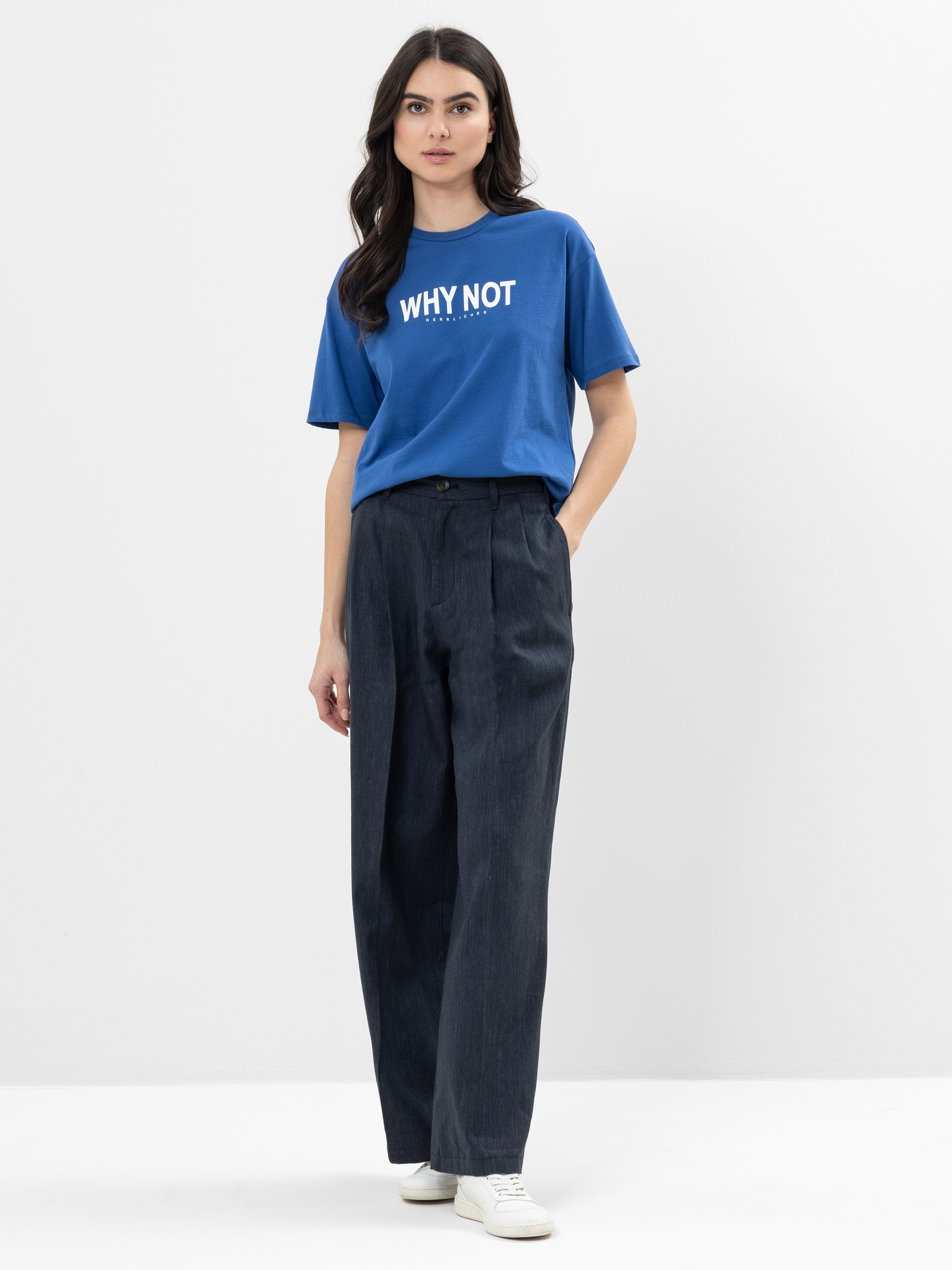Eine Frau steht vor einem schlichten weißen Hintergrund und trägt ein blaues T-Shirt mit der Aufschrift WHY NOT, eine dunkle, weit geschnittene Hose und weiße Turnschuhe. Eine Hand steckt in ihrer Hosentasche und sie schaut mit neutralem Blick in die K
