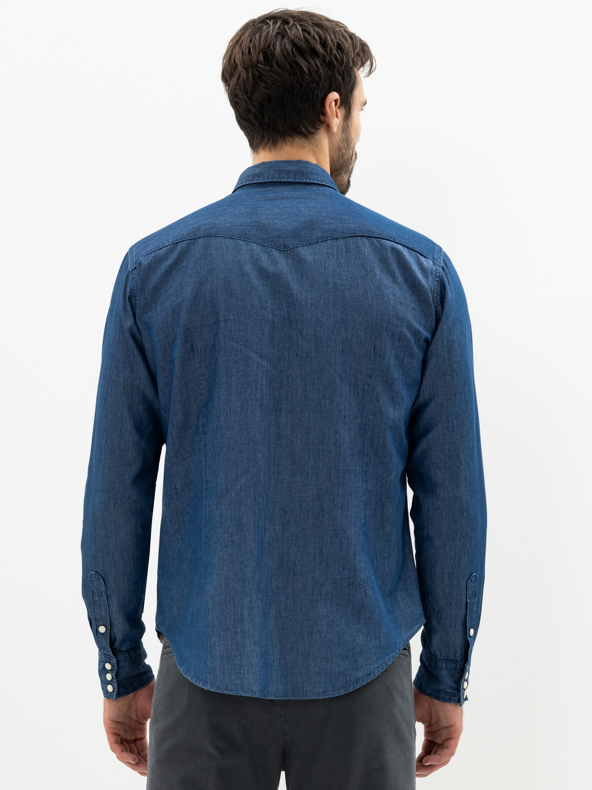 Ein Mann mit kurzen braunen Haaren, von hinten gesehen, trägt ein langärmeliges Jeanshemd und eine dunkle Hose und steht vor einem schlichten weißen Hintergrund.