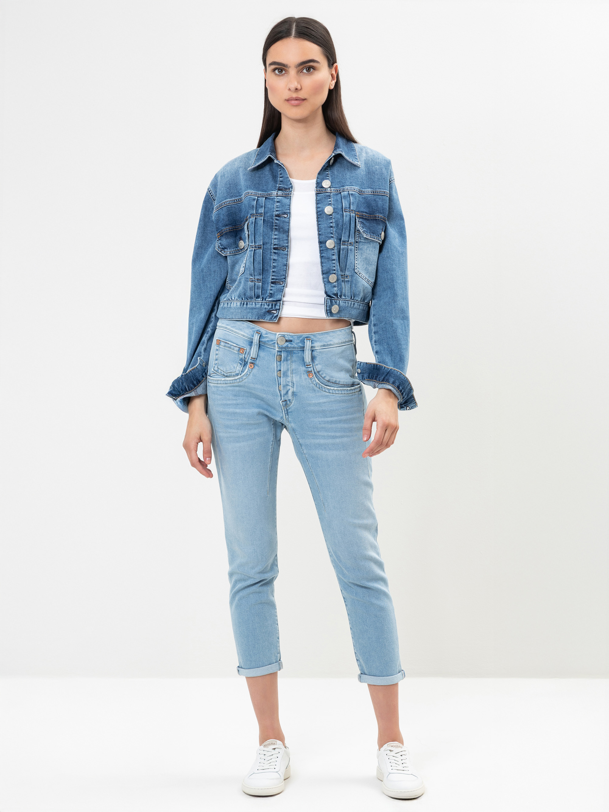 Eine Frau steht vor einem schlichten weißen Hintergrund und trägt eine blaue Jeansjacke, ein weißes Crop-Top, hellblaue Jeans und weiße Turnschuhe. Sie hat langes glattes Haar und einen neutralen Gesichtsausdruck.