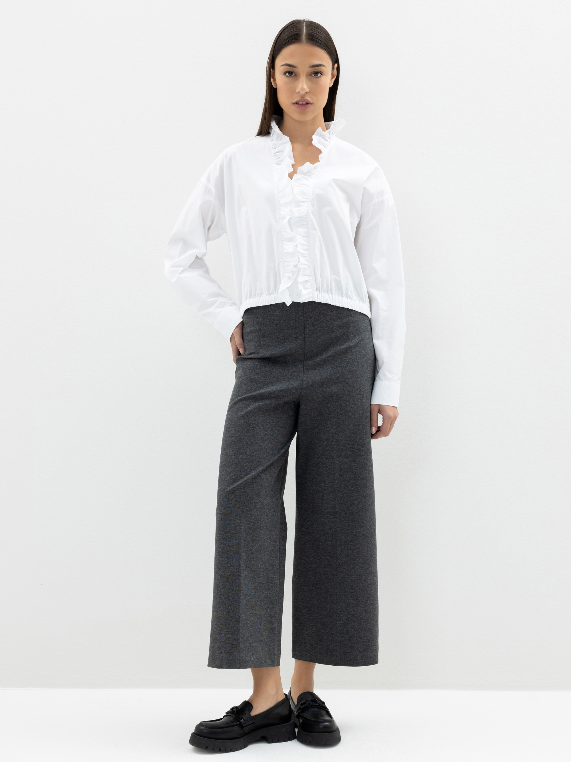 Eine Frau steht vor einem weißen Hintergrund und trägt eine weiße, langärmelige Bluse mit Rüschendetails und eine graue Hose mit hoher Taille und weitem Bein, gepaart mit schwarzen Schuhen. Sie hat glattes, dunkles Haar und einen neutralen Gesichtsau