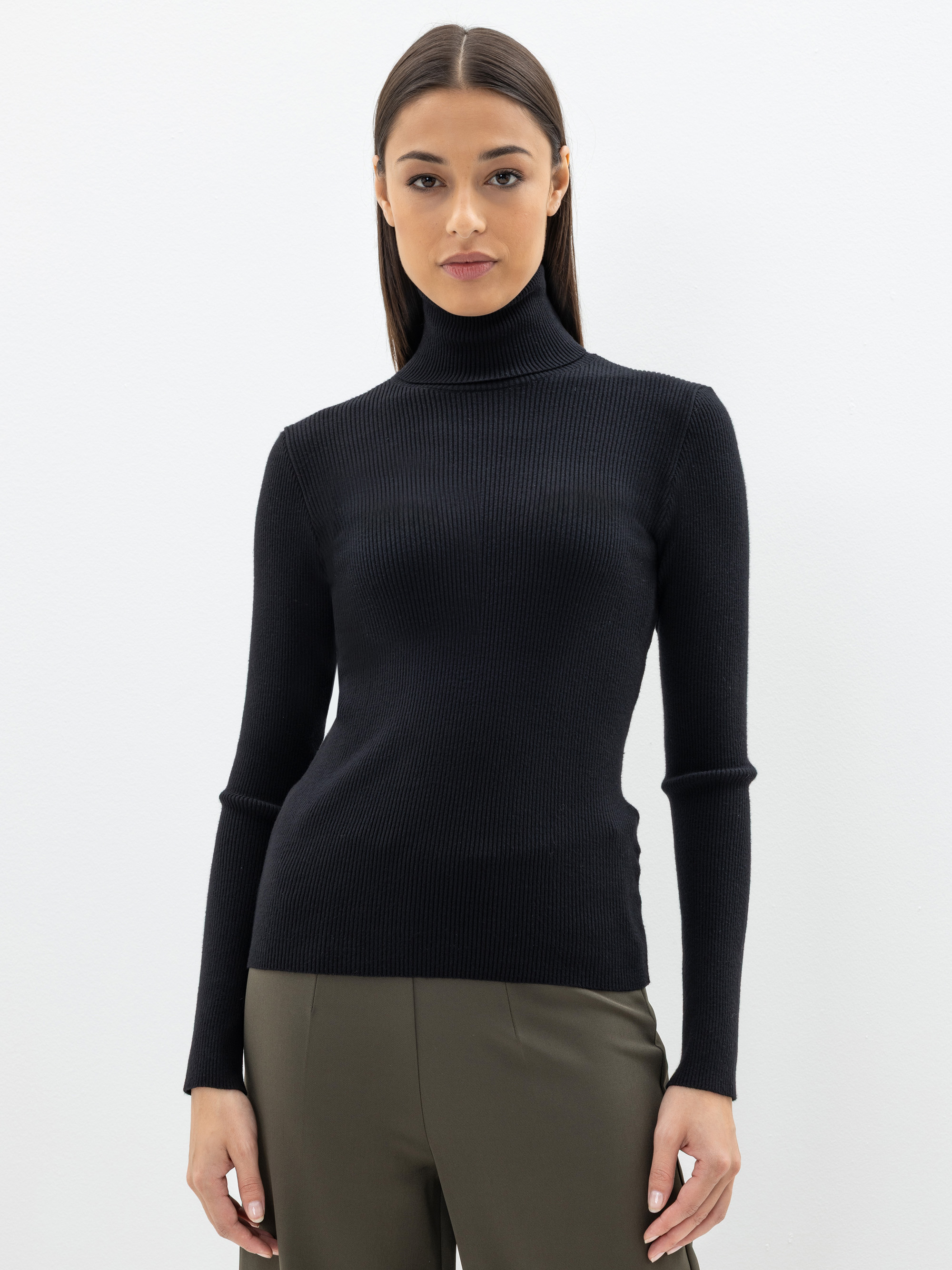 Eine Frau steht vor einem schlichten weißen Hintergrund und trägt einen schwarzen Rollkragenpullover und eine olivgrüne Hose. Sie hat glattes braunes Haar und einen neutralen Gesichtsausdruck, die Arme liegen entspannt an ihren Seiten.