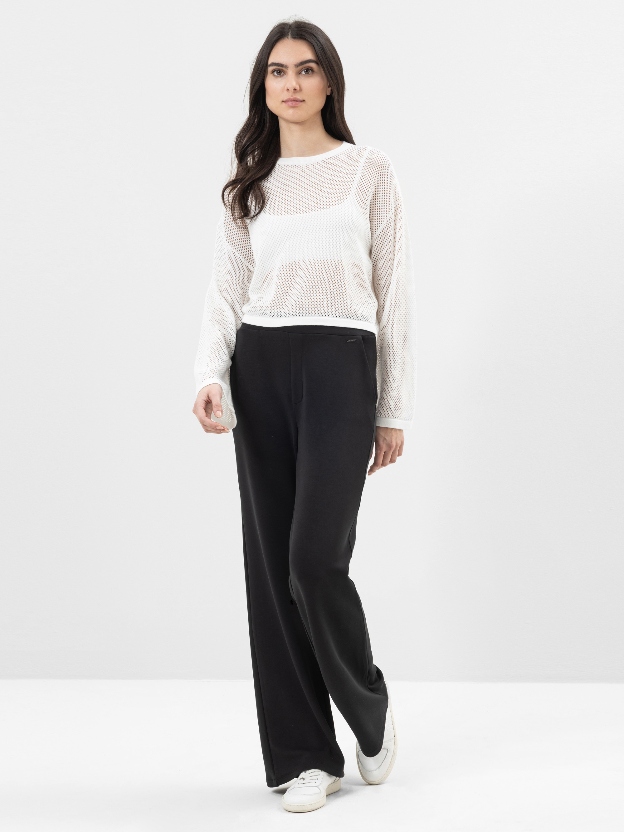 Eine Frau steht vor einem schlichten weißen Hintergrund und trägt ein weißes, langärmeliges Oberteil über einem weißen Tank, eine schwarze Hose mit weitem Bein und weiße Turnschuhe. Sie hat langes dunkles Haar und einen neutralen Gesichtsausdruck.