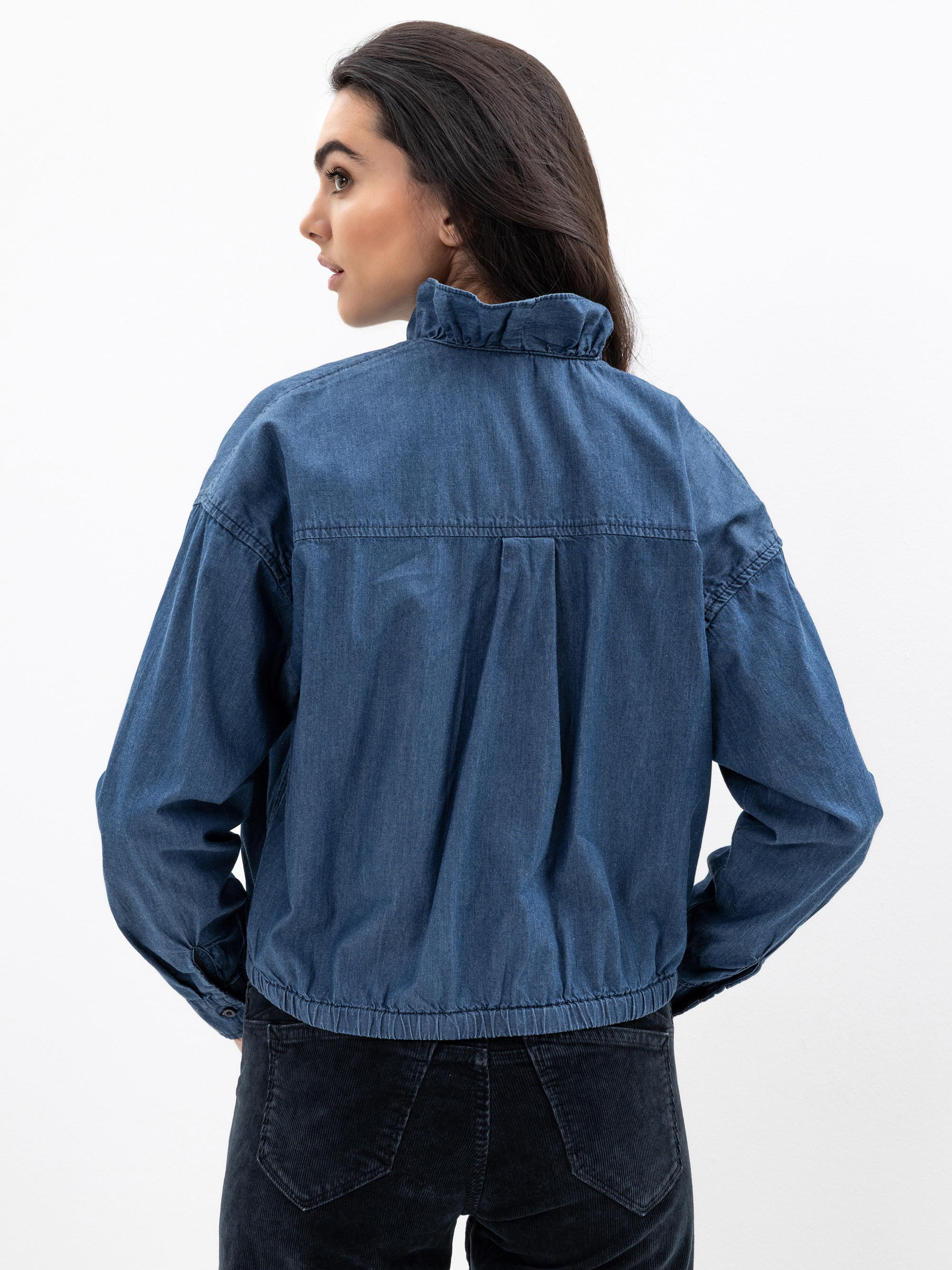 Eine Frau mit langen dunklen Haaren, von hinten gesehen, trägt eine blaue Jeansjacke mit hohem Kragen und schwarze Jeans. Sie blickt nach links vor einem schlichten weißen Hintergrund.