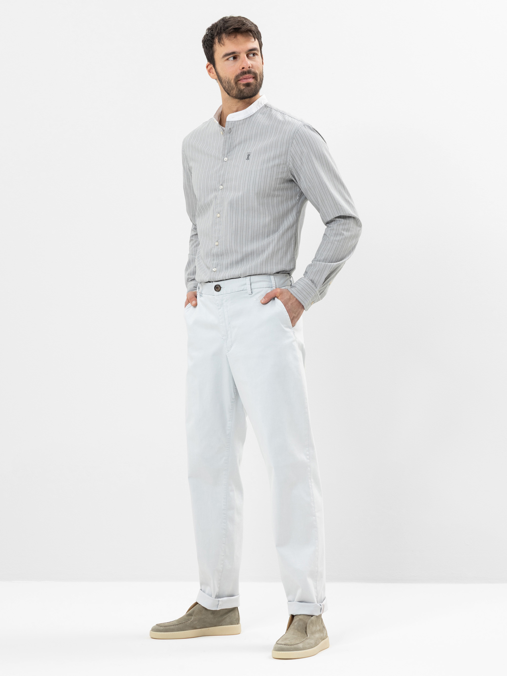 Ein Mann steht vor einem schlichten weißen Hintergrund, trägt ein hellgrau gestreiftes Button-up-Hemd, eine helle Hose und beigefarbene Wildlederschuhe. Er hat die Hände in den Taschen und blickt zur Seite.