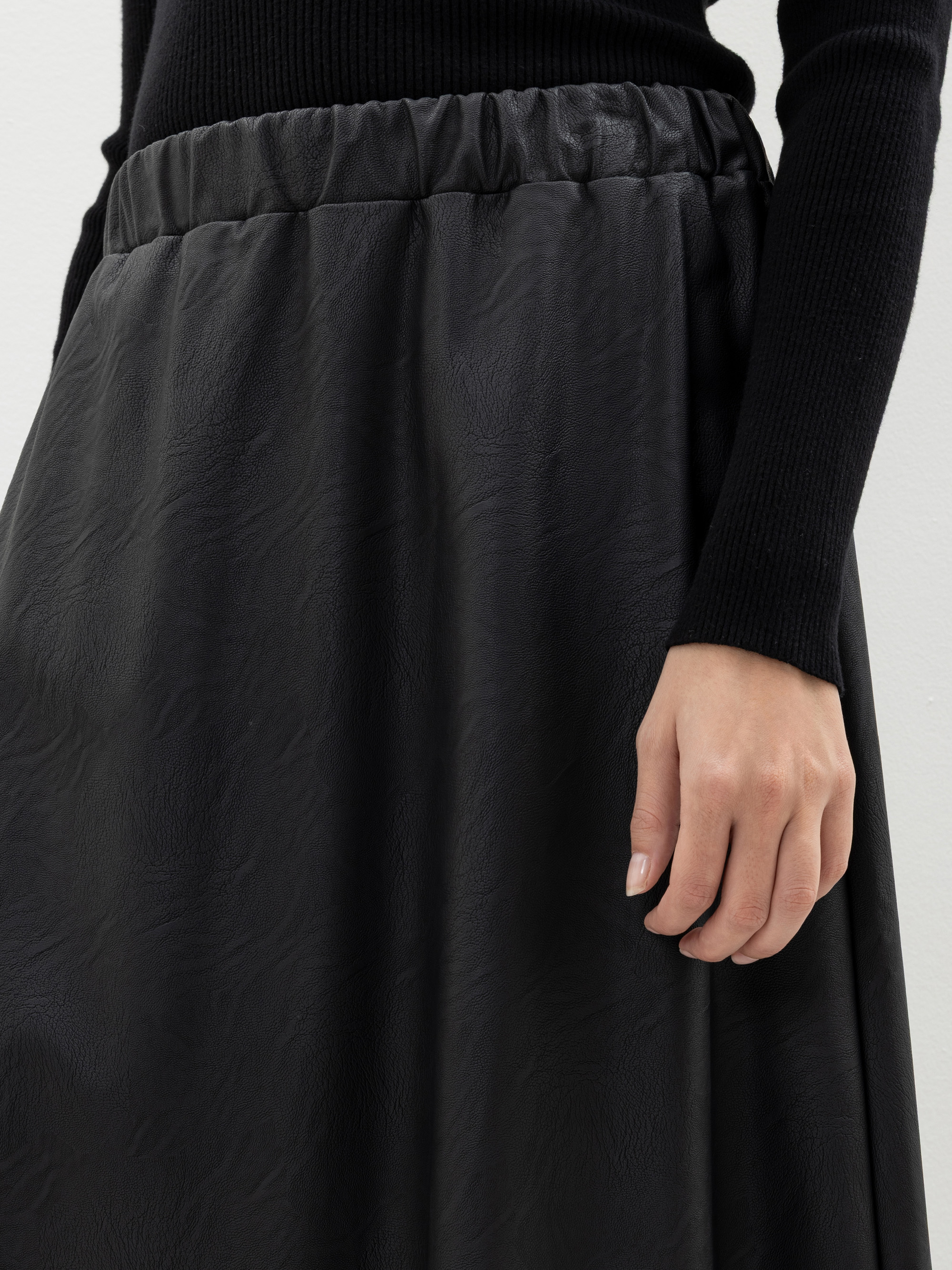 Eine Person, die einen langen schwarzen Rock mit einem strukturierten Muster und einem elastischen Bund trägt, gepaart mit einem schwarzen gerippten Pullover. Nur der untere Teil des Oberkörpers und die rechte Hand sind sichtbar.