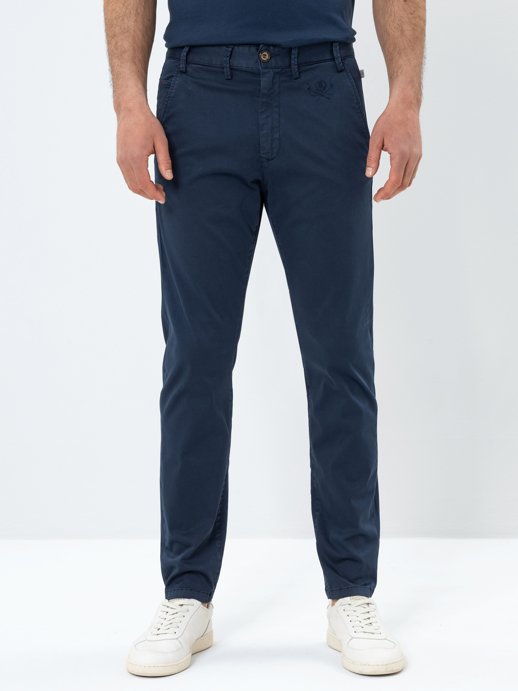 Eine Person, die eine marineblaue Chinohose, ein passendes marineblaues Hemd und weiße Turnschuhe trägt, steht vor einem schlichten weißen Hintergrund. Das Bild konzentriert sich auf die Hose und zeigt sie von der Taille bis zu den Knöcheln.