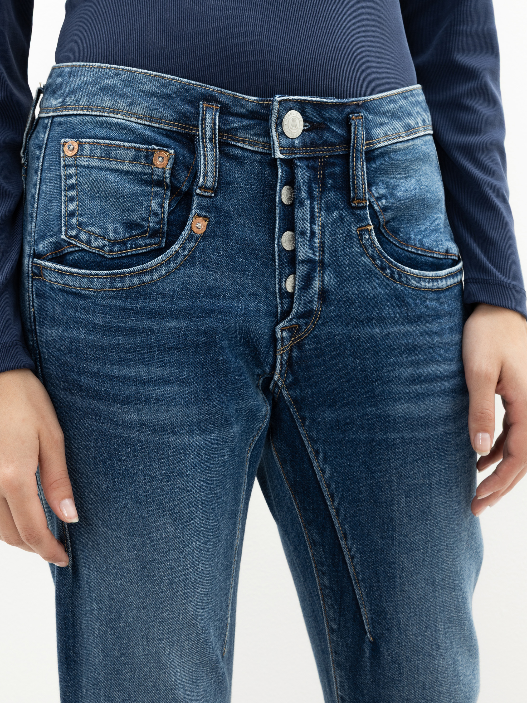 Eine Person, die eine blaue Jeans mit teilweise geöffnetem Hosenschlitz, der silberne Knöpfe freigibt, und ein marineblaues langärmeliges Oberteil trägt. Nur der Oberkörper und die Oberschenkel sind sichtbar.
