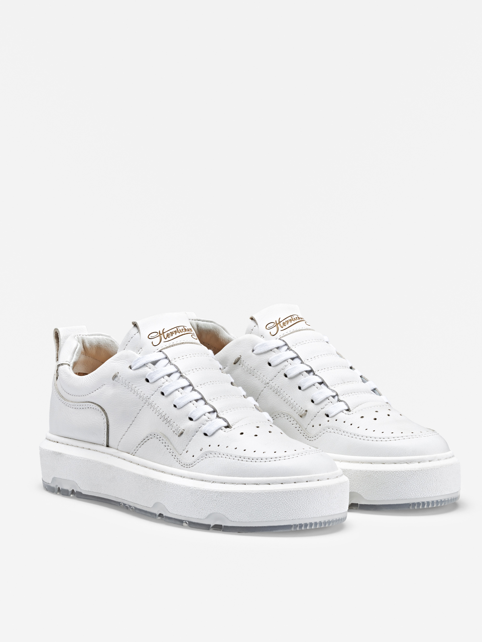 Ein Paar weiße Ledersneaker mit perforierten Details, Schnürung vorne und dicker weißer Sohle, die vor einem einfarbig weißen Hintergrund stehen.