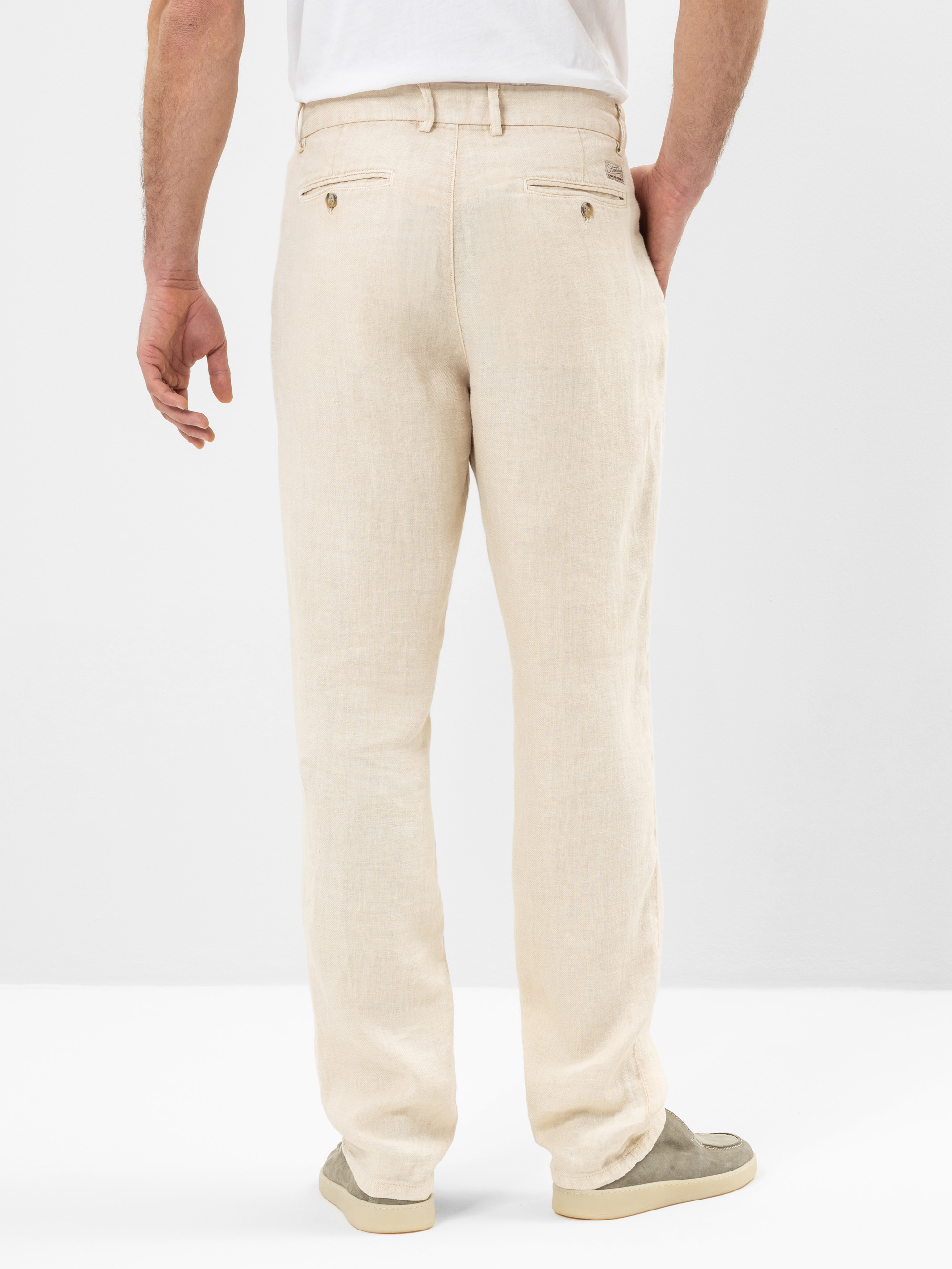 Eine Person, die eine hellbeige Leinenhose, ein weißes Hemd und hellgraue Schuhe trägt, wird von hinten gezeigt und steht vor einem schlichten weißen Hintergrund.