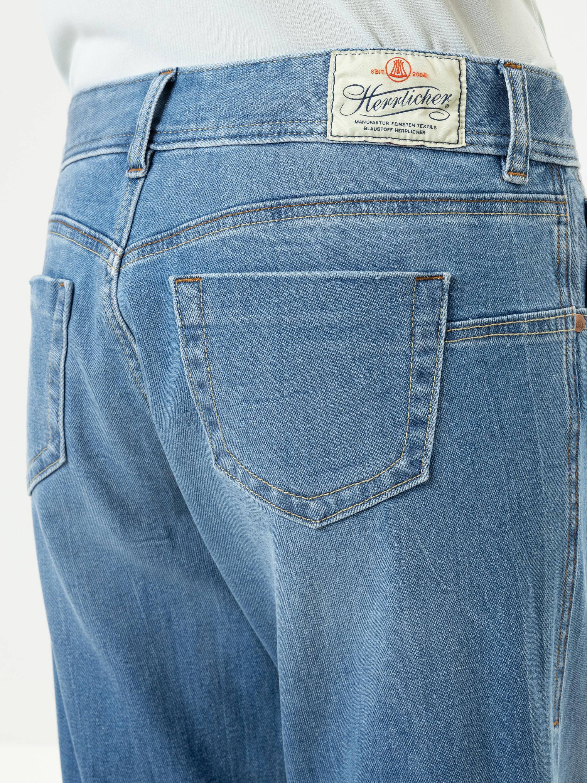 Nahaufnahme des Rückens einer Person, die eine hellblaue Denim-Jeans mit einem weißen Herrlicher-Markenaufnäher über der rechten Tasche trägt. Die Jeans hat klassische Nähte und eine verblasste Waschung.
