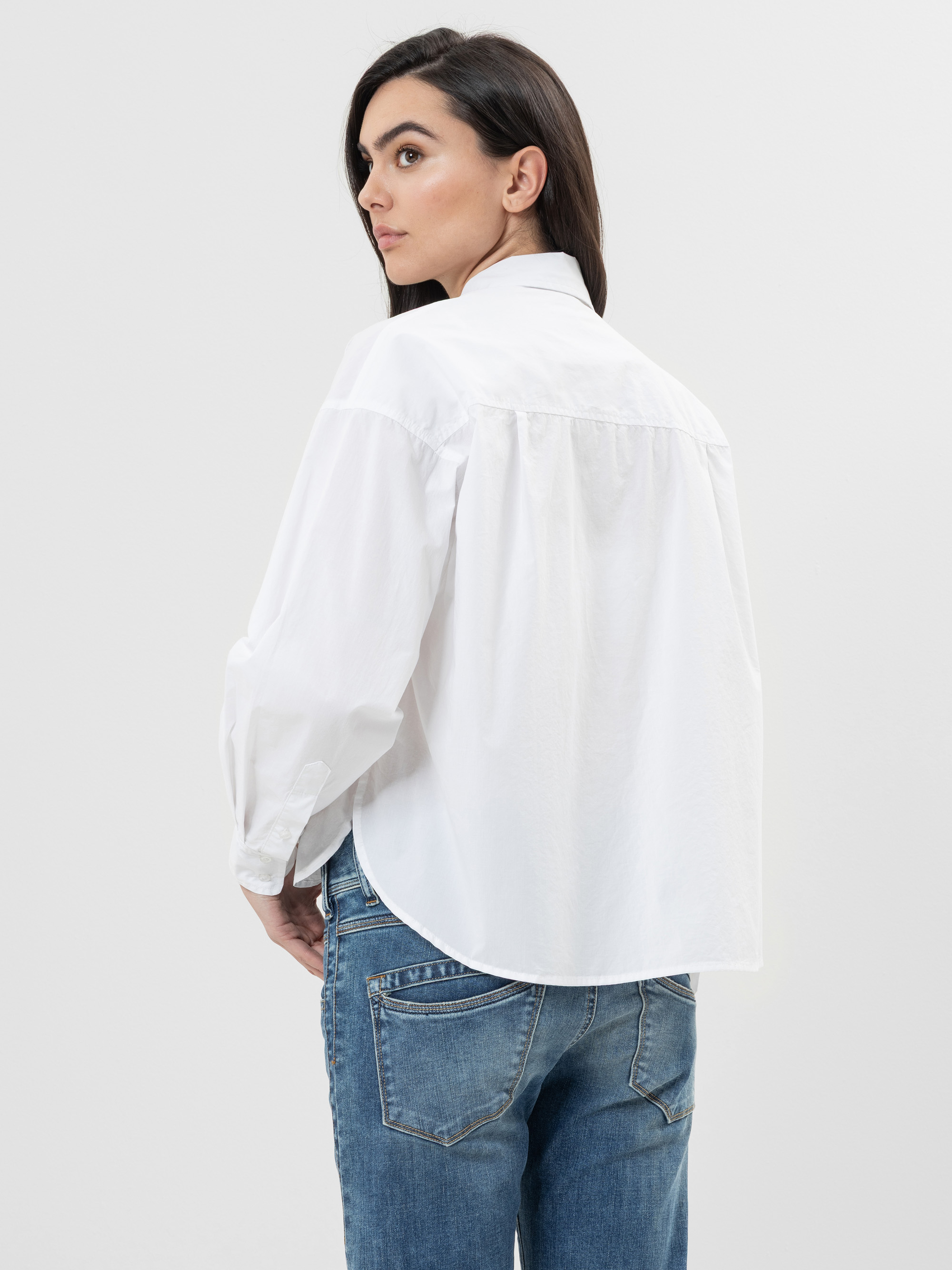 Eine Frau mit langen dunklen Haaren, die ein lockeres weißes Button-up-Hemd und blaue Jeans trägt, steht mit dem Gesicht zur Kamera und blickt über ihre Schulter vor einem schlichten hellen Hintergrund zurück.
