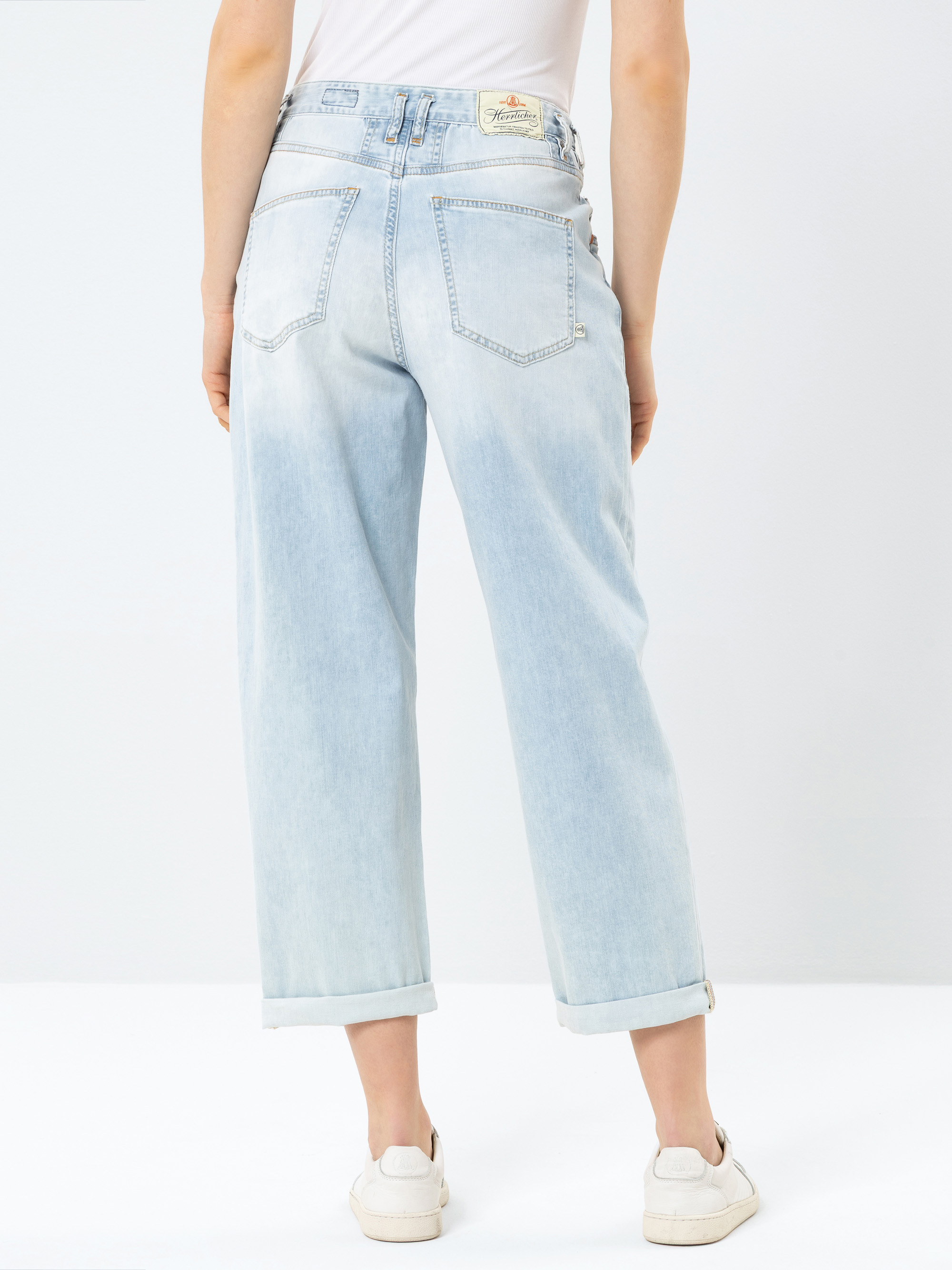 Eine Person, die eine hellblaue Jeans mit hoher Taille und aufgerollten Manschetten, ein weißes Oberteil und weiße Turnschuhe trägt, wird von hinten vor einem schlichten hellen Hintergrund gezeigt.