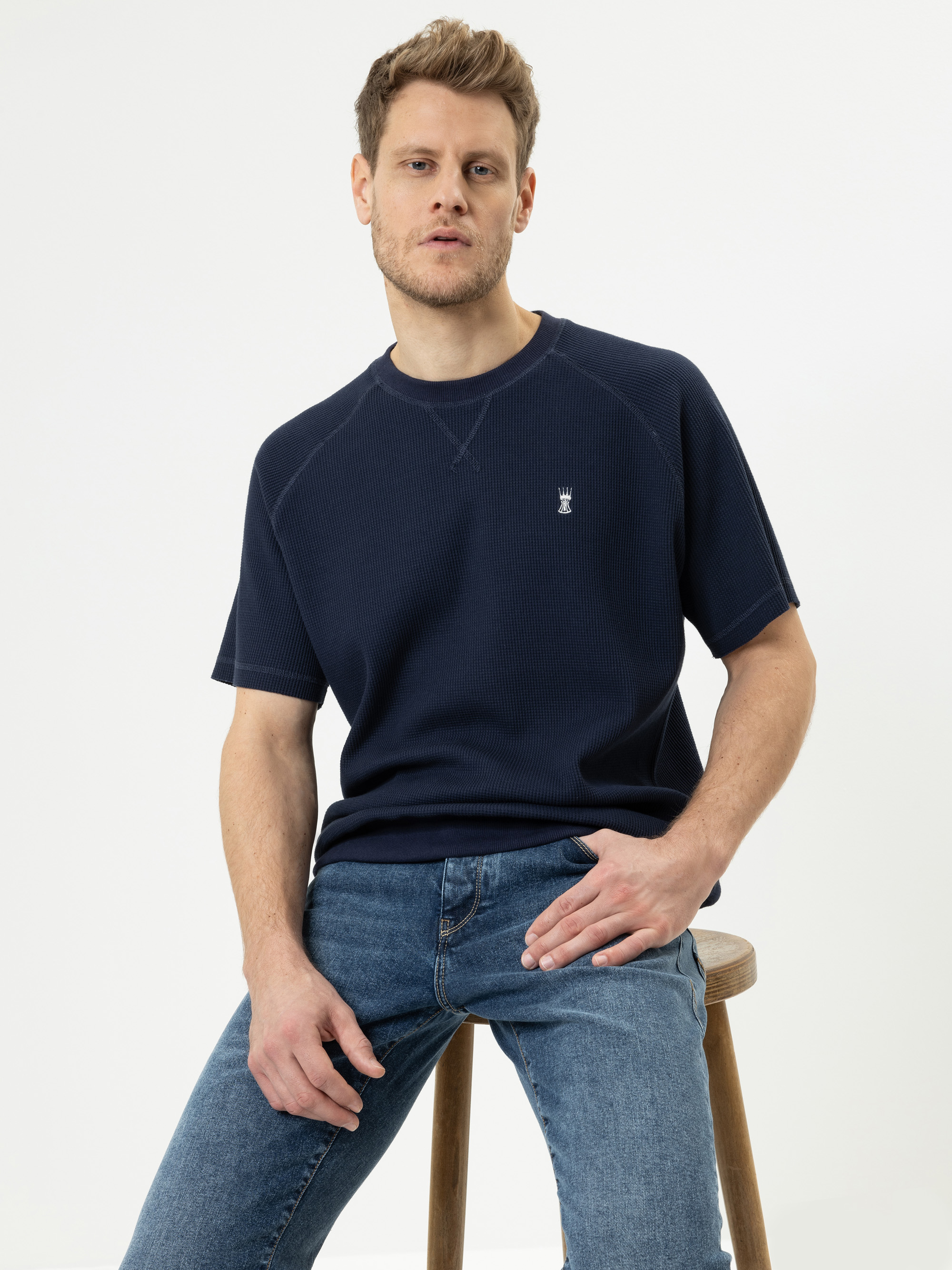 Ein Mann mit hellbraunem Haar, der ein marineblaues kurzärmeliges Hemd und blaue Jeans trägt, sitzt auf einem Holzhocker vor einem schlichten weißen Hintergrund und blickt mit neutralem Blick in die Kamera.