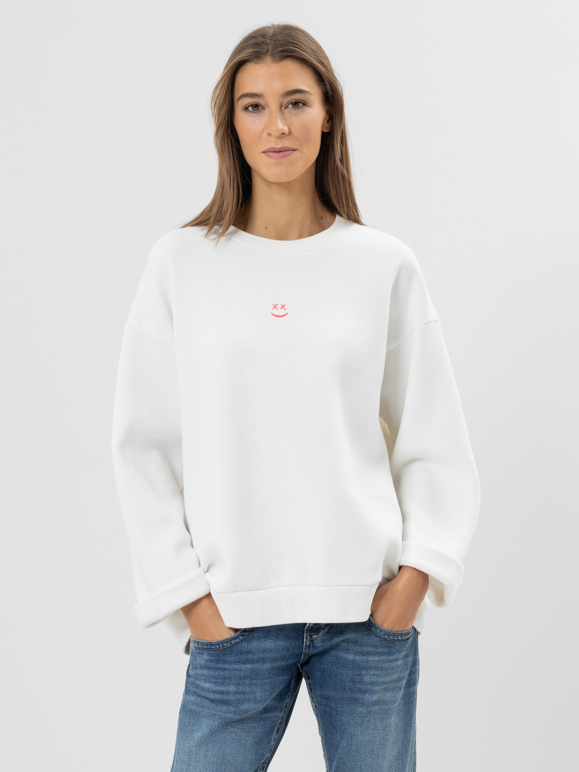 Eine Frau steht vor einem schlichten Hintergrund und trägt ein locker sitzendes weißes Sweatshirt mit einem kleinen rosa Smiley auf der Vorderseite und blaue Jeans. Ihre Hände stecken in den Taschen und sie lächelt sanft in die Kamera.