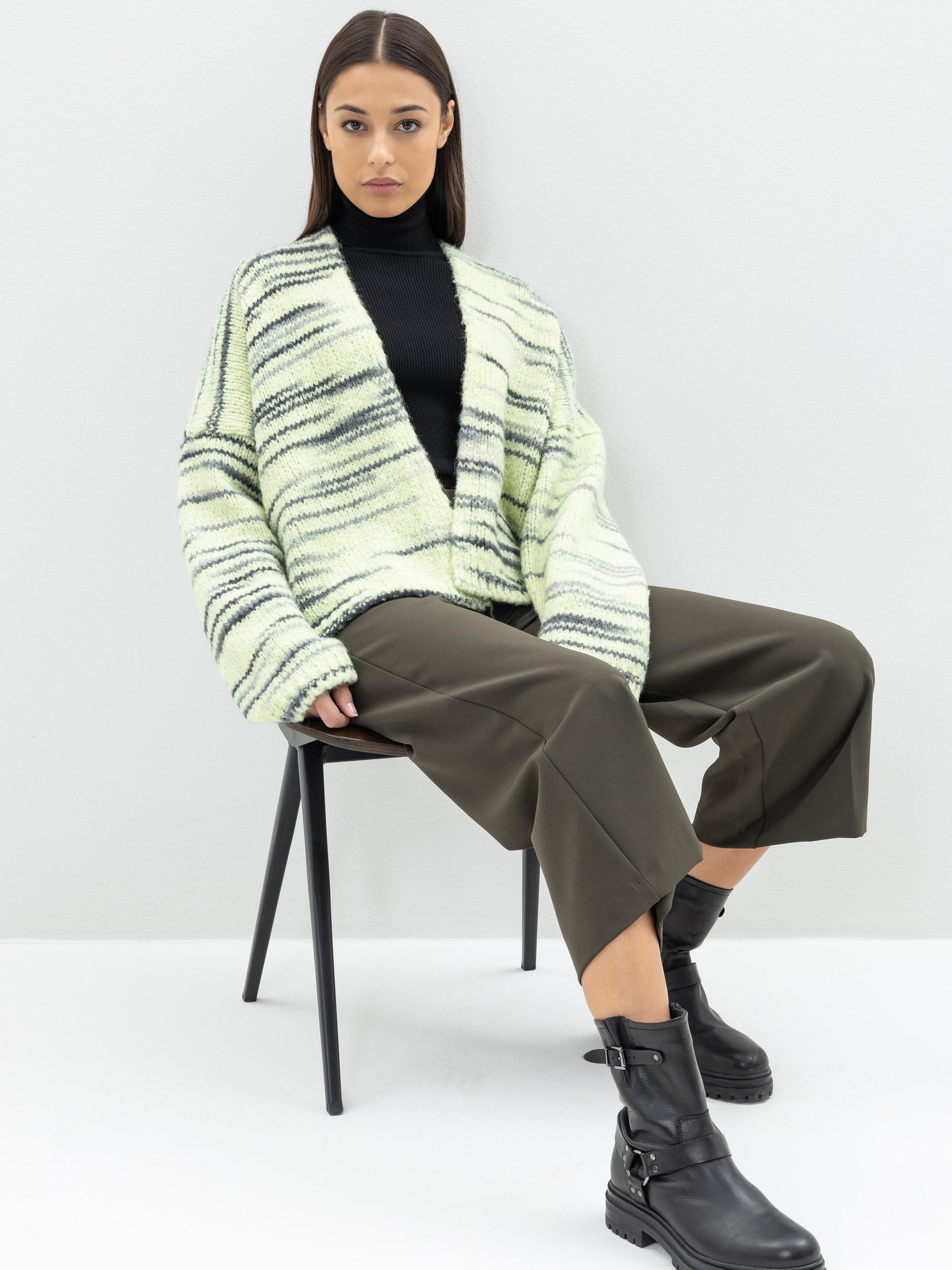 Eine Frau sitzt auf einem Stuhl und trägt einen schwarzen Rollkragenpullover, eine übergroße gestreifte Strickjacke, eine weite olivgrüne Hose und schwarze Stiefel vor einem schlichten hellen Hintergrund.