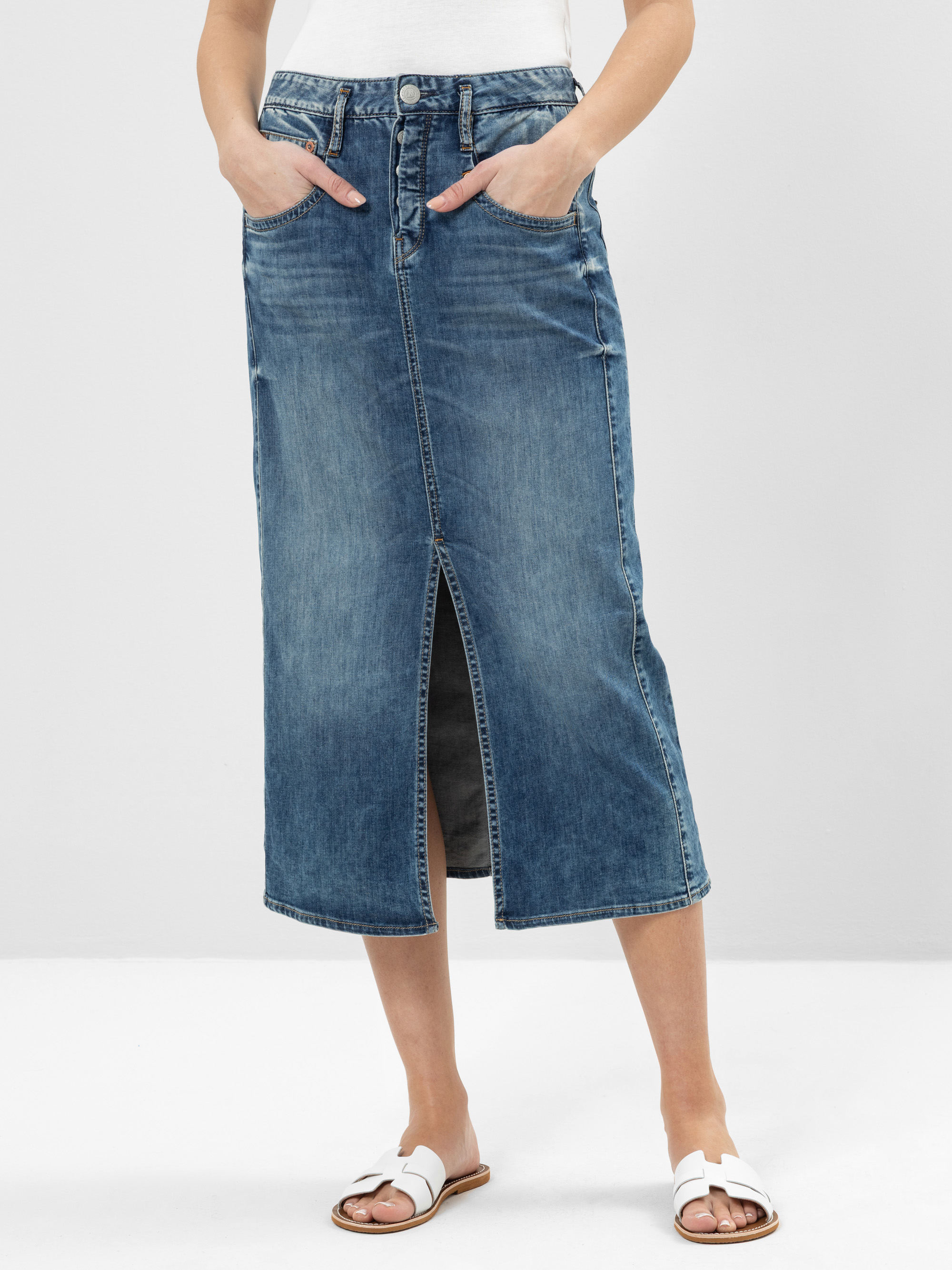 Eine Frau, die einen blauen Jeans-Midirock mit Schlitz, ein weißes Oberteil und weiße Sandalen trägt, posiert mit den Händen in den Rocktaschen vor einem schlichten hellen Hintergrund.