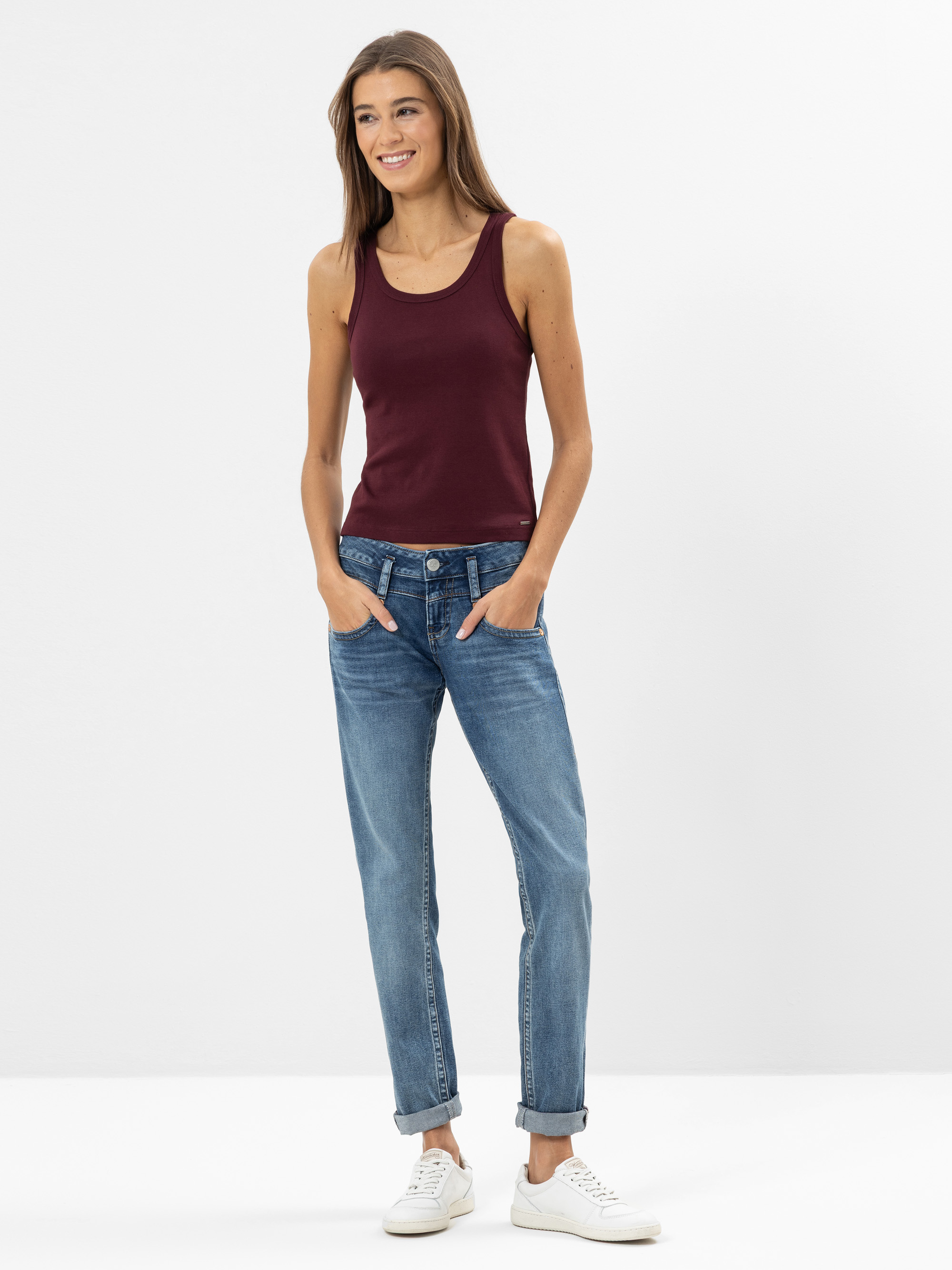 Eine Frau steht vor einem schlichten weißen Hintergrund und trägt ein weinrotes Tanktop, blaue Jeans mit aufgerollten Bündchen und weiße Turnschuhe. Sie hat langes braunes Haar und lächelt mit den Händen in den Taschen.