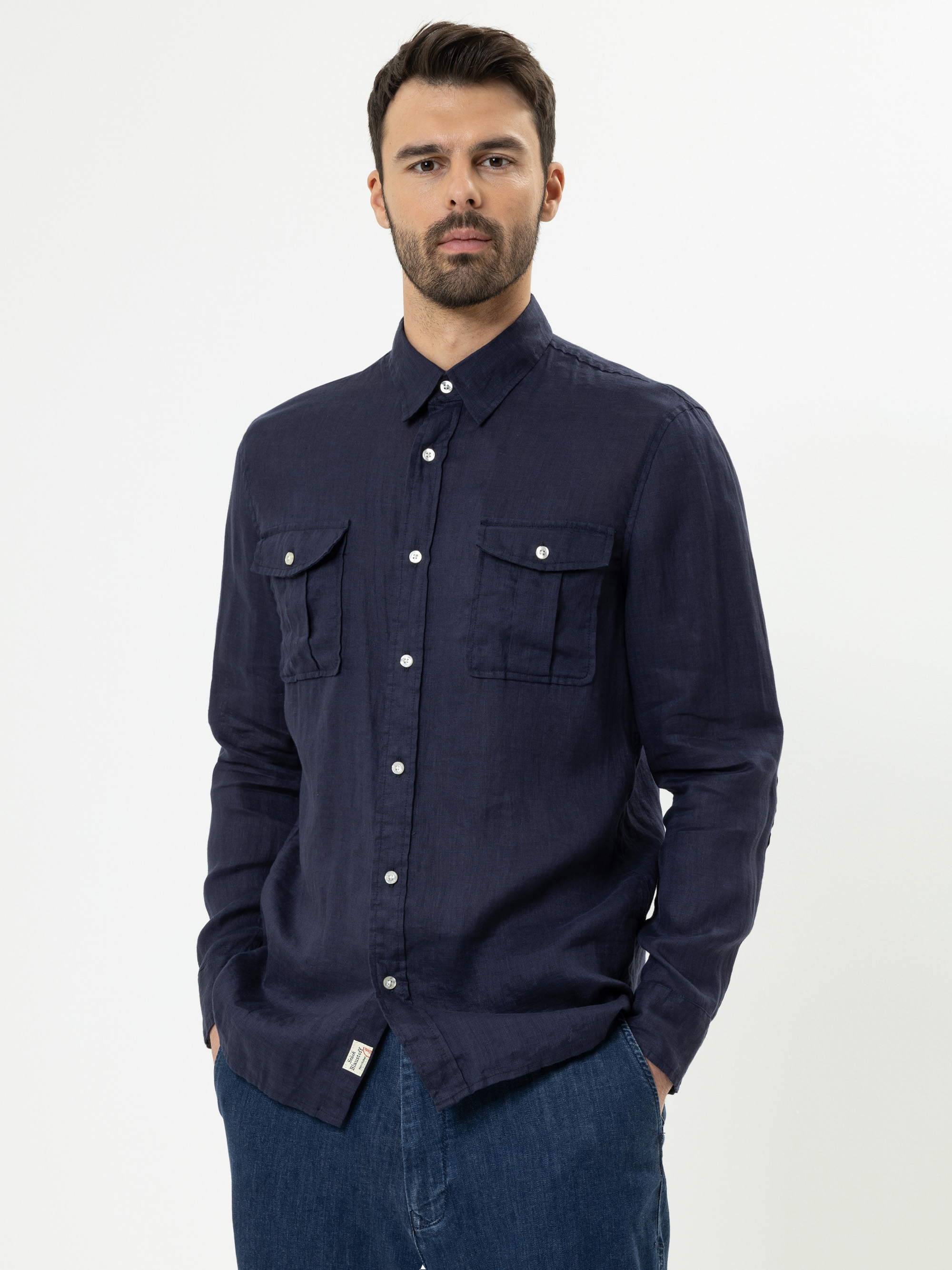 Ein Mann mit dunklem Haar und Bart trägt ein marineblaues Button-up-Hemd mit zwei Brusttaschen und blaue Jeans und steht mit den Händen in den Taschen vor einem schlichten weißen Hintergrund.