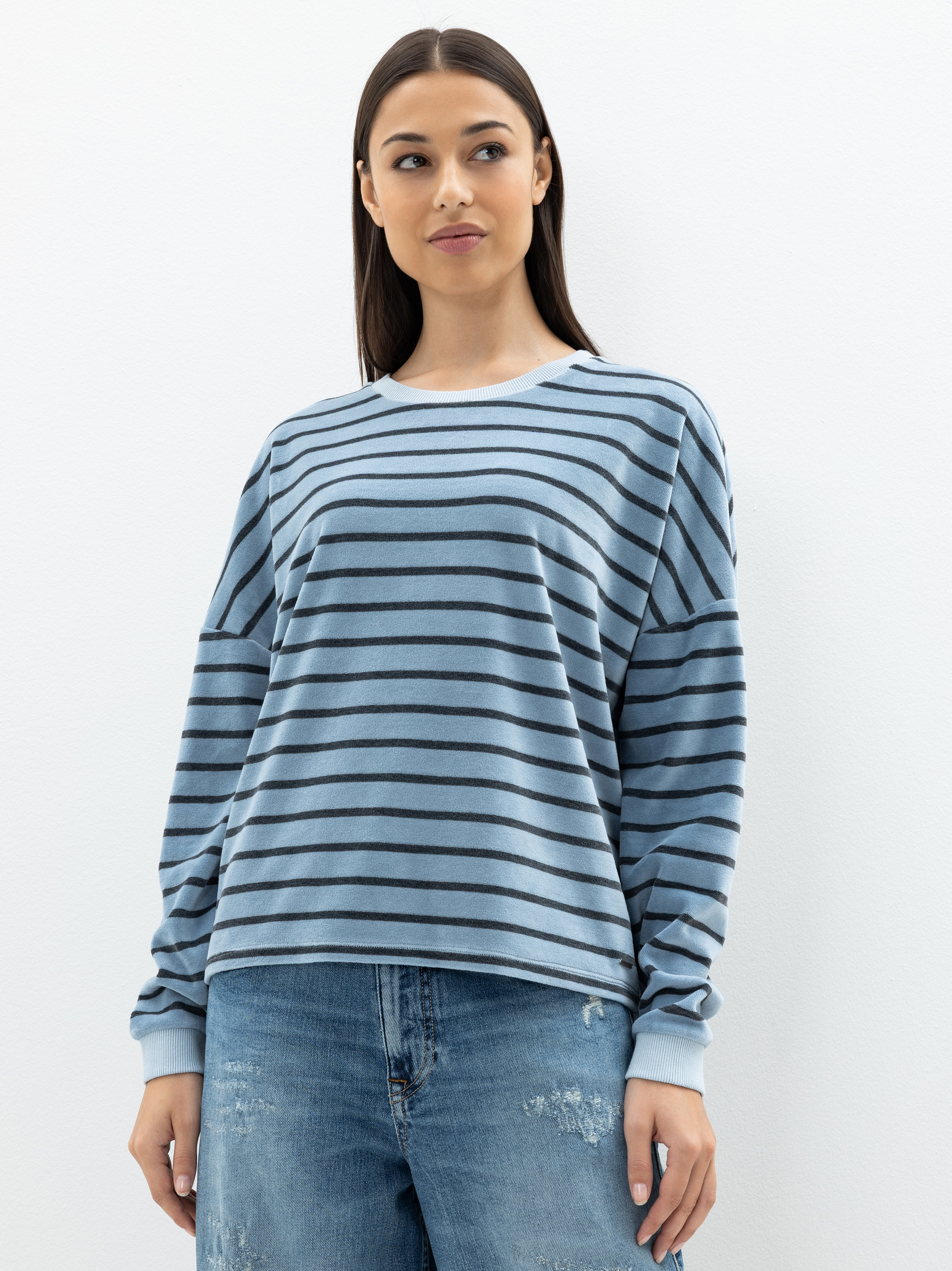 Eine Frau mit langen dunklen Haaren trägt ein hellblau-schwarz gestreiftes langärmeliges Hemd und verblichene blaue Jeans und steht vor einem schlichten weißen Hintergrund.