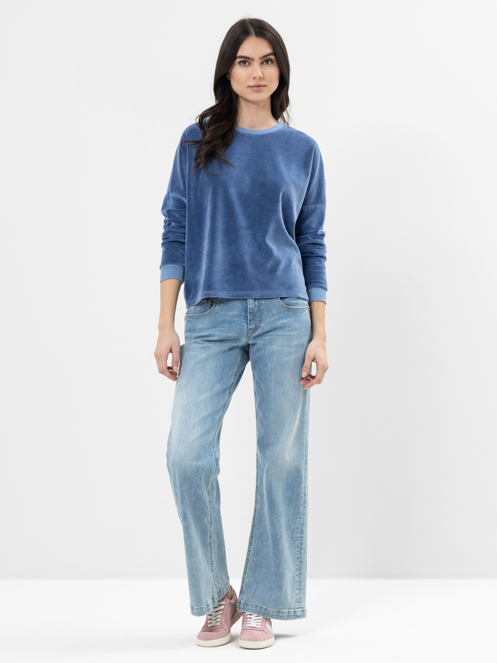 Eine Frau steht vor einem schlichten weißen Hintergrund und trägt einen blauen langärmeligen Pullover, hellblaue Jeans mit weitem Bein und rosa Turnschuhe. Sie hat langes dunkles Haar und einen neutralen Gesichtsausdruck.